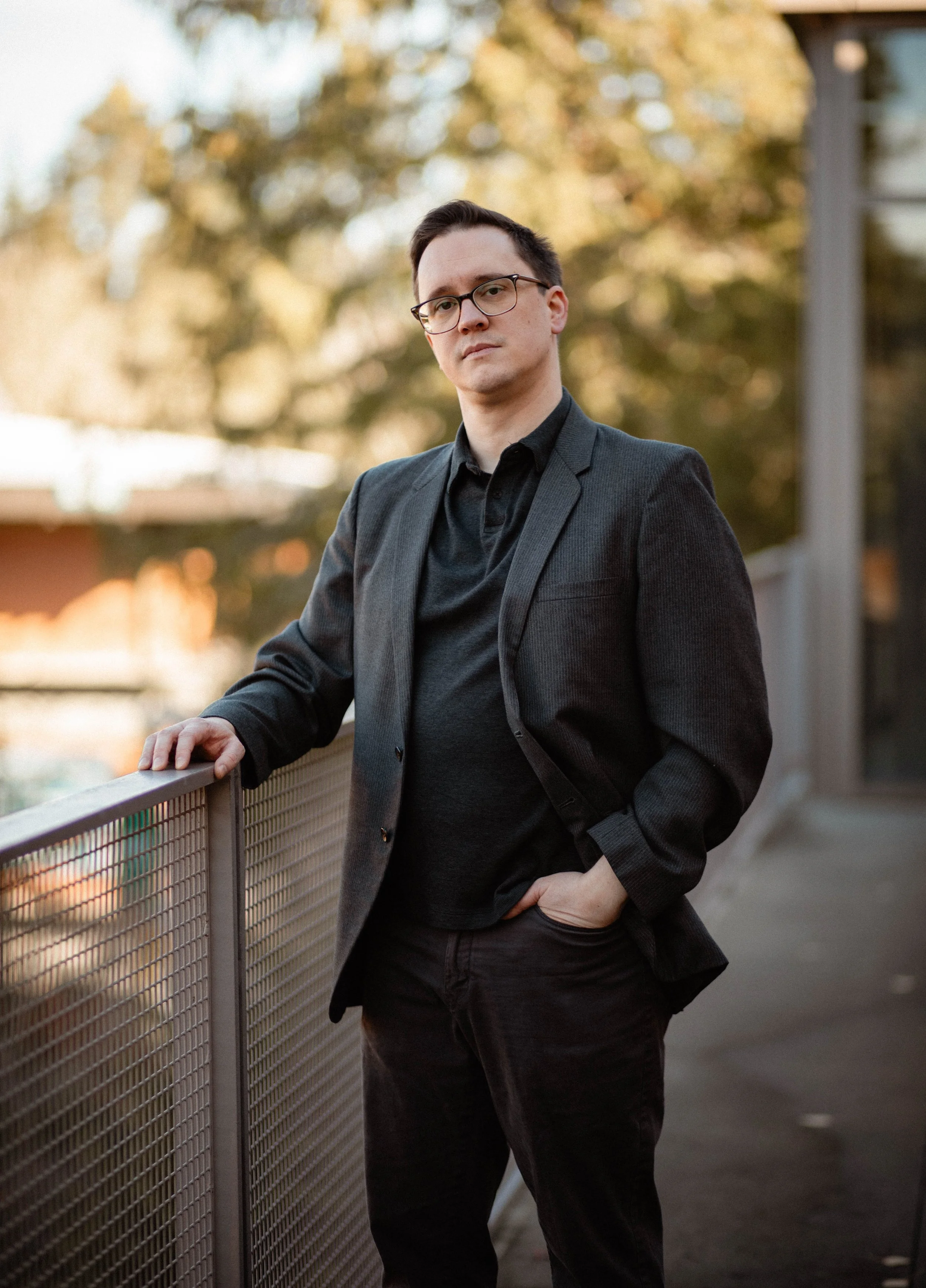 A man wearing glasses, a black shirt, and a dark blazer standing outdoors near a metal railing, with trees and a building in the background during late afternoon or early evening. Seattle professional head shot photography