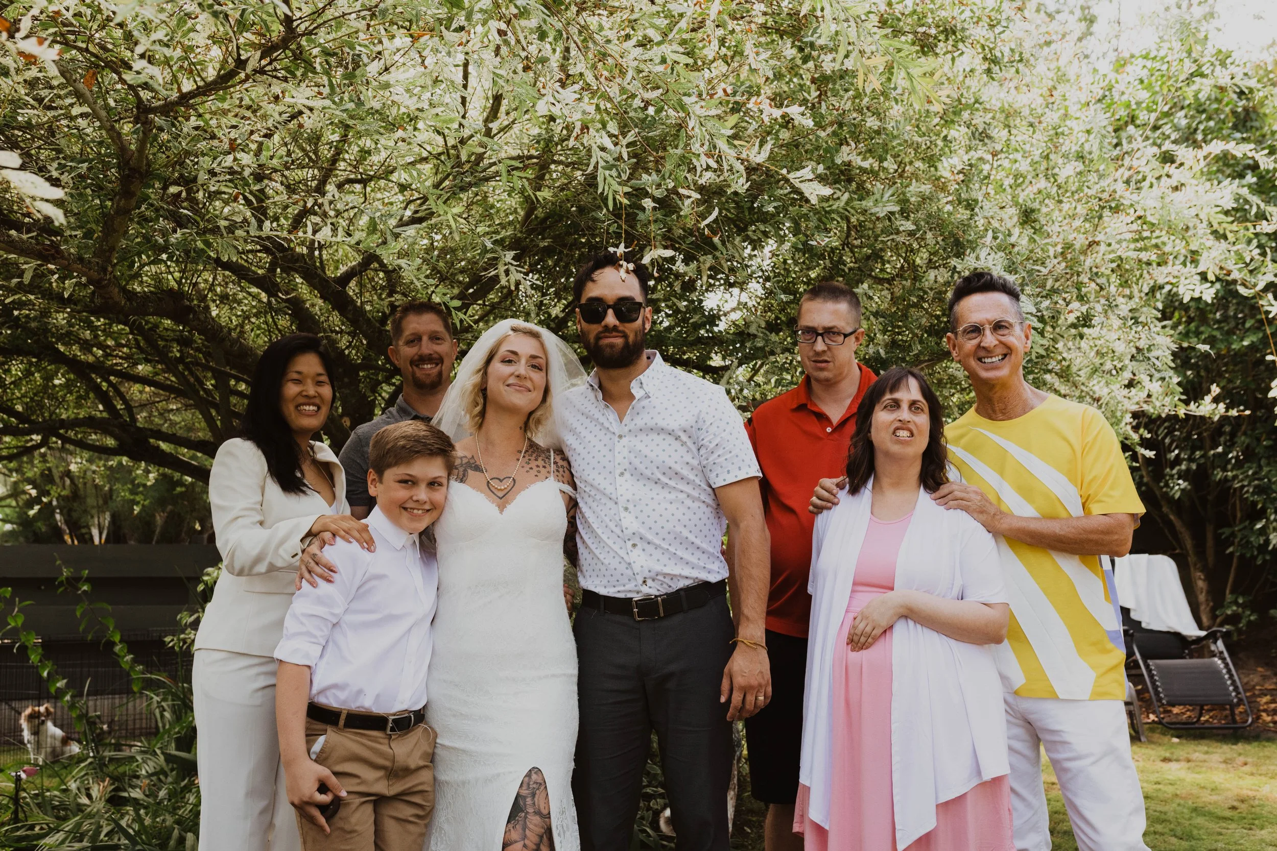 Group of eight people standing outdoors under a tree, celebrating a wedding. The bride, wearing a lace wedding dress and veil, is in the center. The groom, in a white shirt and sunglasses, is beside her. Others are in casual and semi-formal attire, s