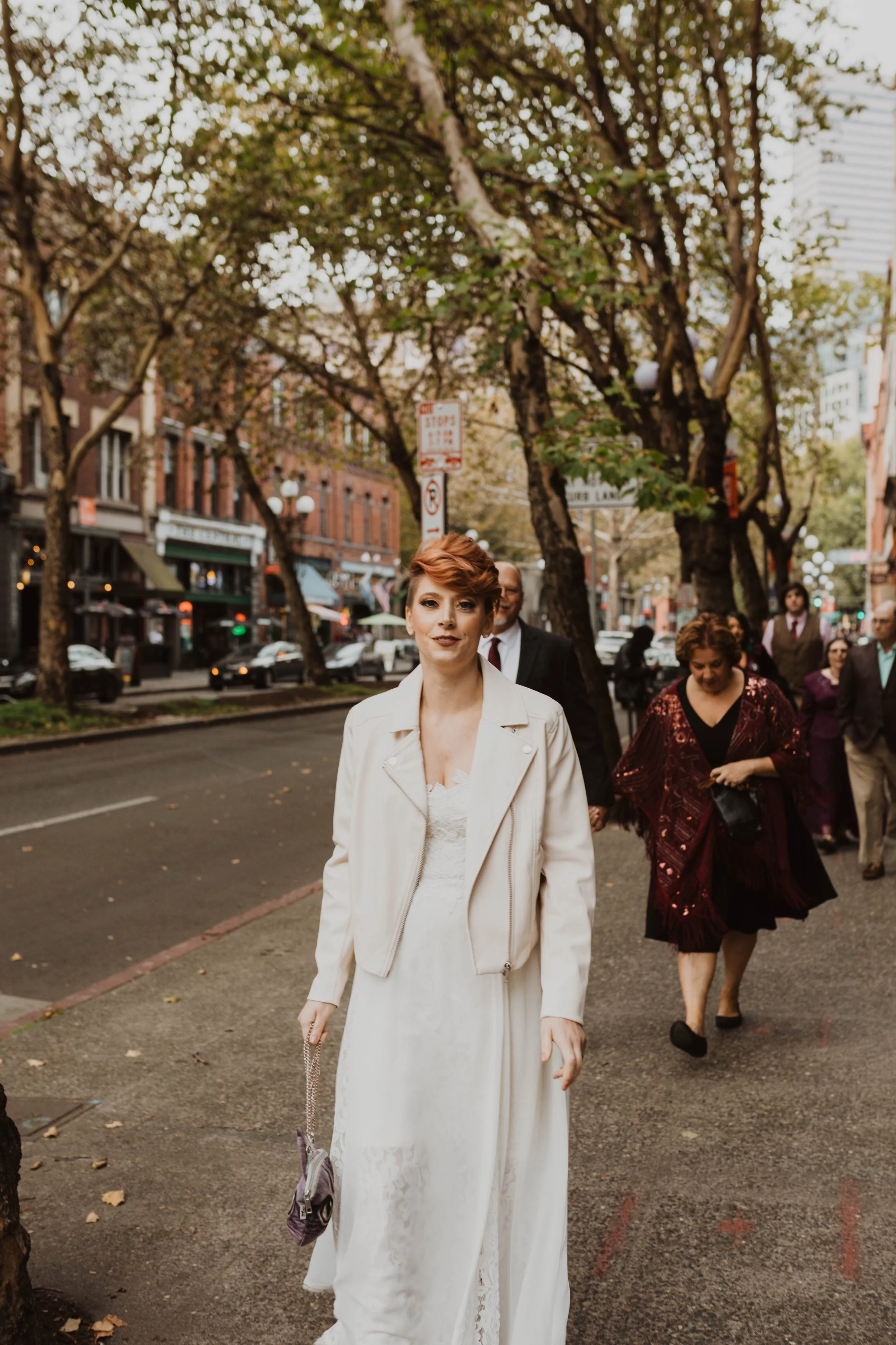 A woman in a white dress and jacket walking on a city sidewalk, surrounded by other pedestrians and trees with autumn leaves. Pioneer Square, Seattle, WA wedding photography.