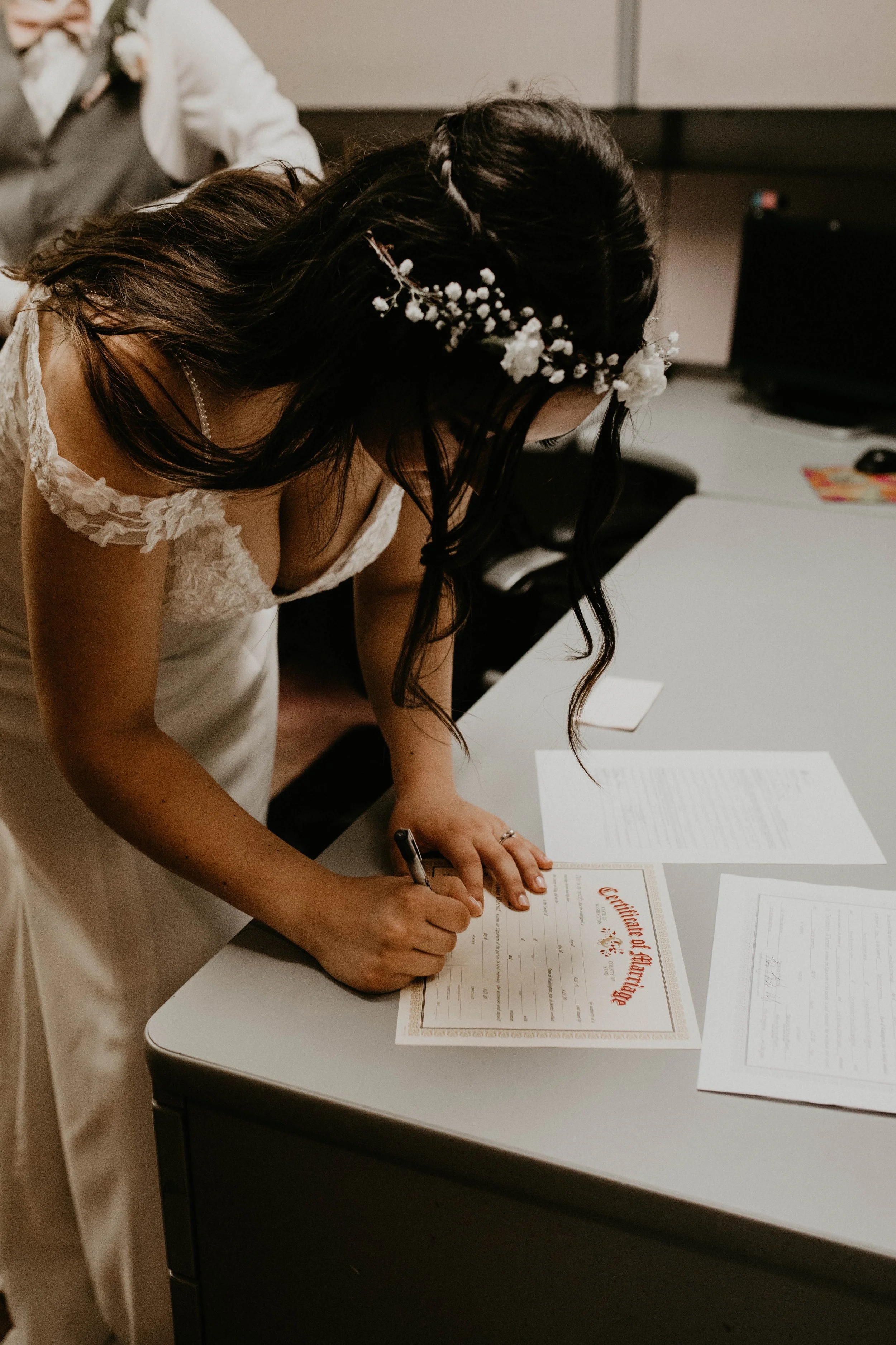 Bridal bride signing wedding certificate at a table, wearing a white dress with lace details and a floral headband, with documents and papers spread out on the table. Seattle, WA wedding photography.