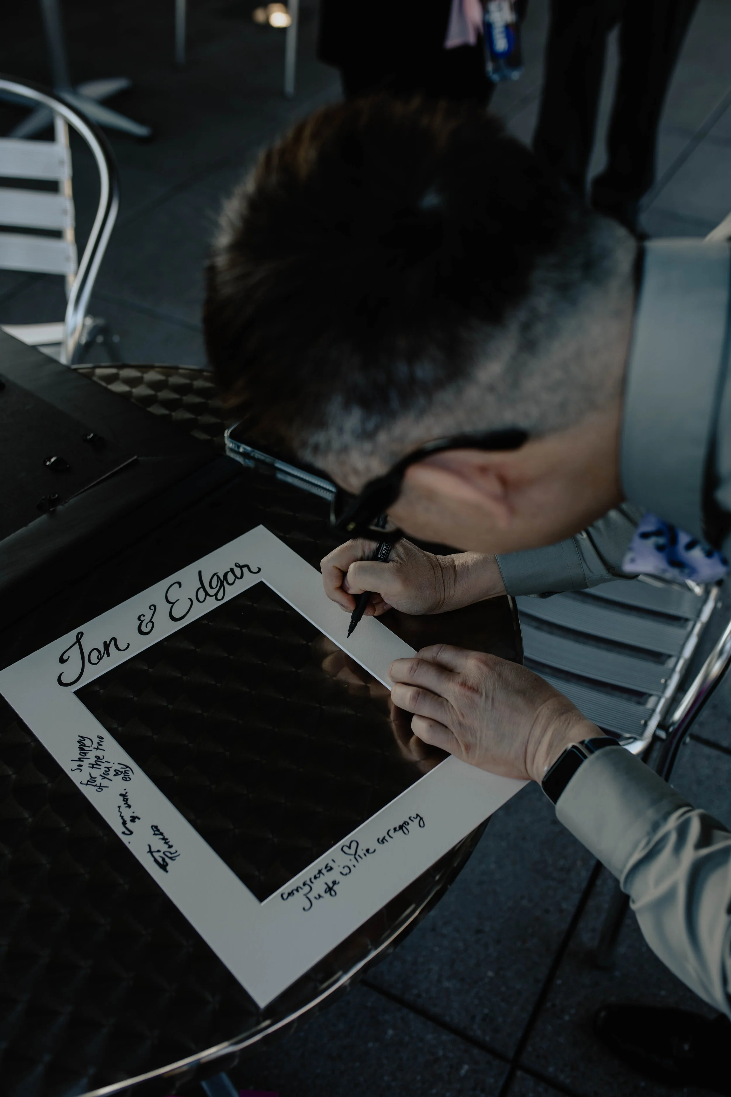 A man in a gray suit is writing on a white picture frame with black handwritten text at a table, with the names 'Jan & Edgar' at the top and various messages and signatures inside. Seattle Municipal Courthouse wedding photography.