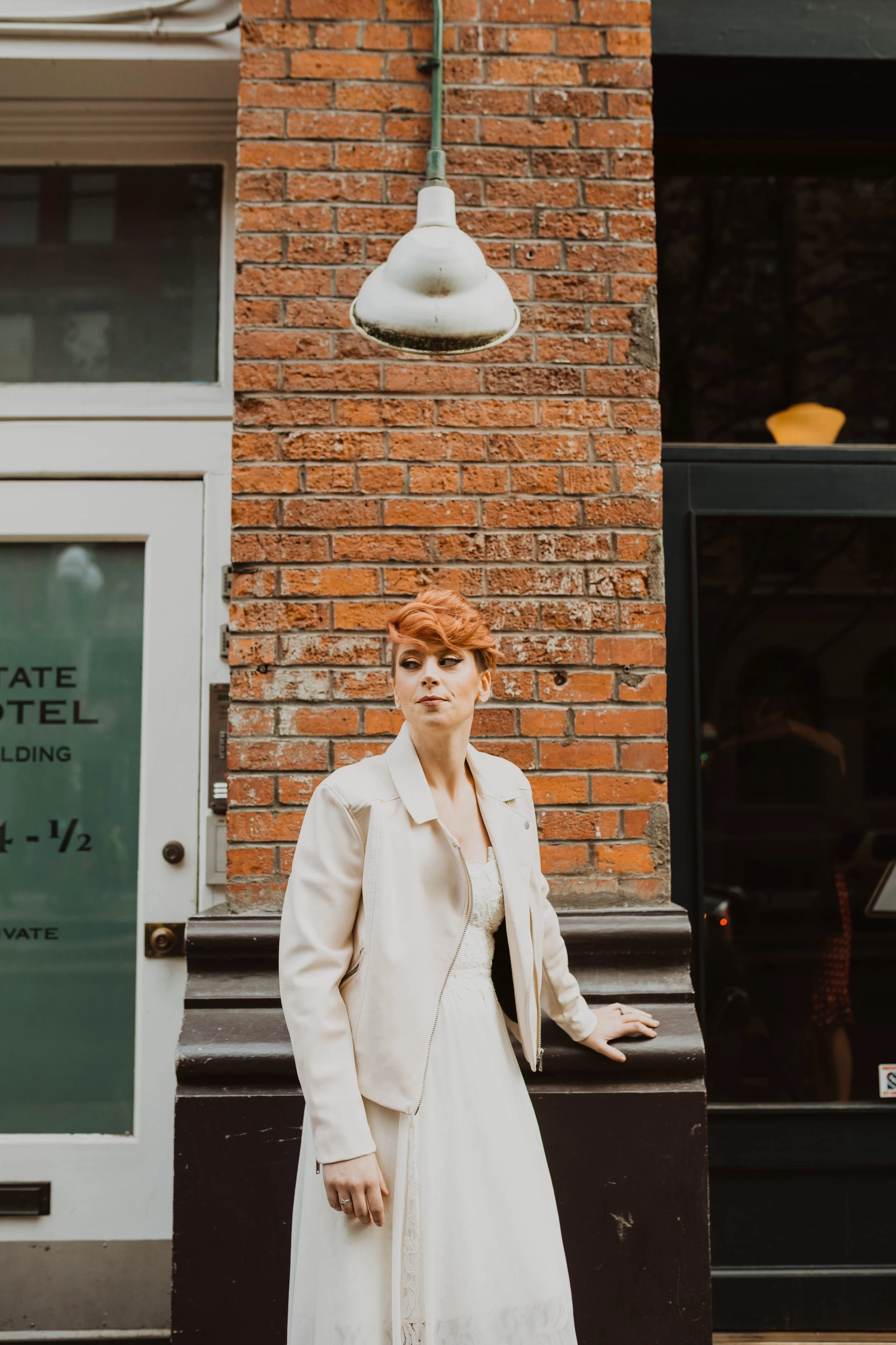 A woman with short red hair wearing a white dress and cream jacket, standing against a red brick building exterior, with one hand resting on a black ledge and looking to the side. Pioneer Square, Seattle, WA wedding photography.
