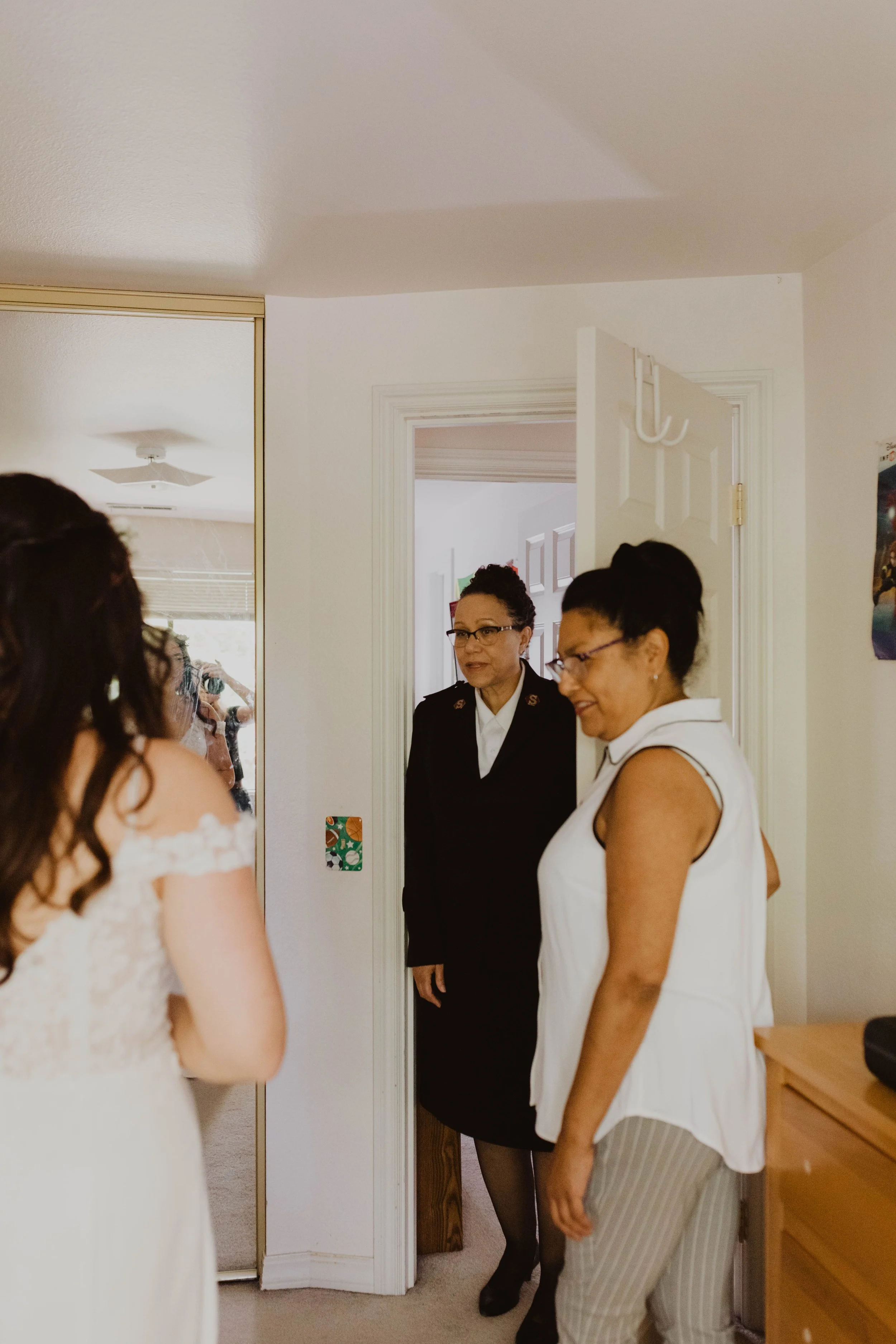 Three women are in a room, with one woman partially visible on the left, a woman in a black uniform in the center, and a woman wearing glasses and a white vest on the right. They are standing close together near a doorway, and a mirror reflects a per