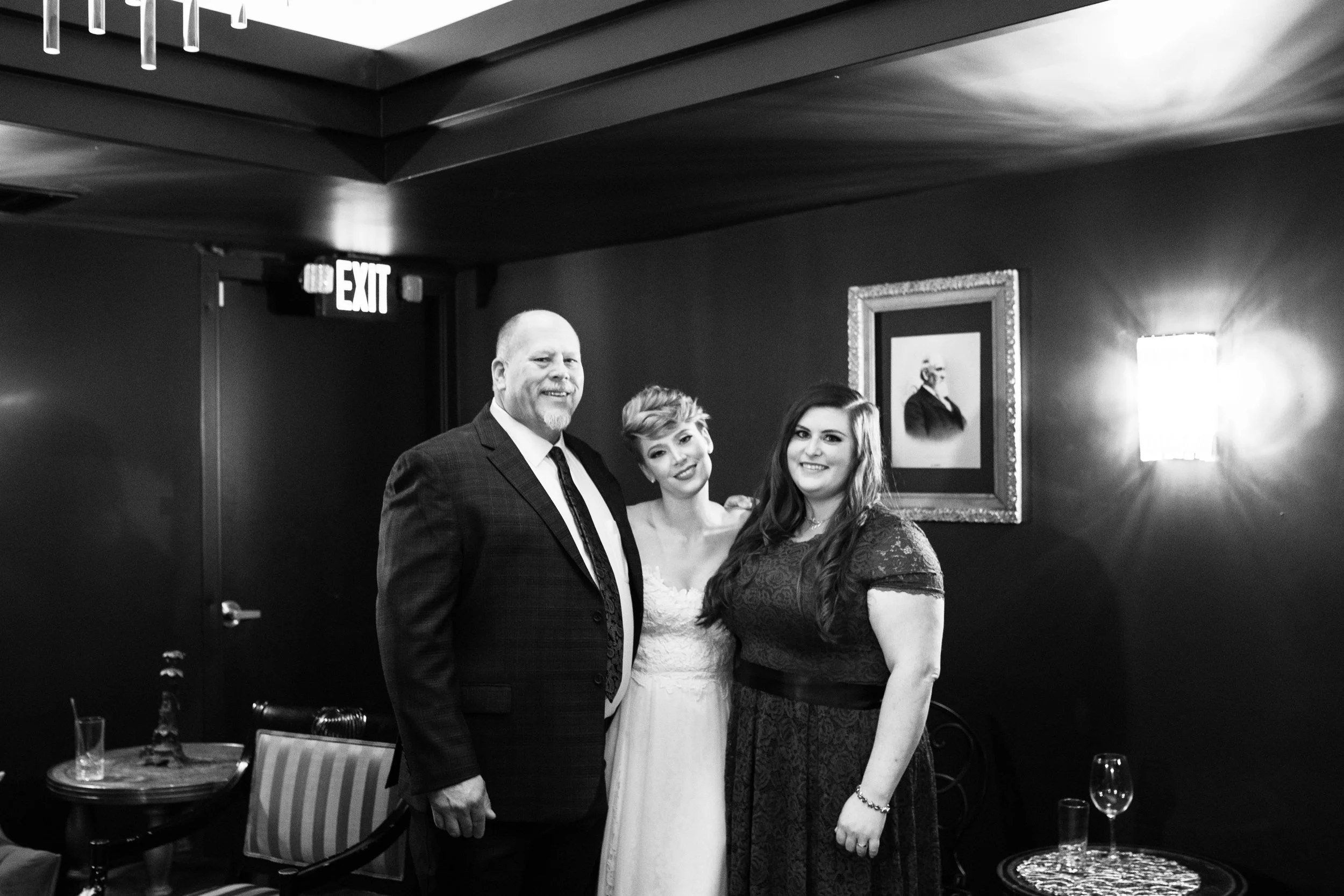 A black-and-white photo of three people smiling indoors. The man on the left has a beard and is wearing a suit, the woman in the middle is in a white dress, and the woman on the right has long dark hair and is wearing a dark dress. They stand in a ro