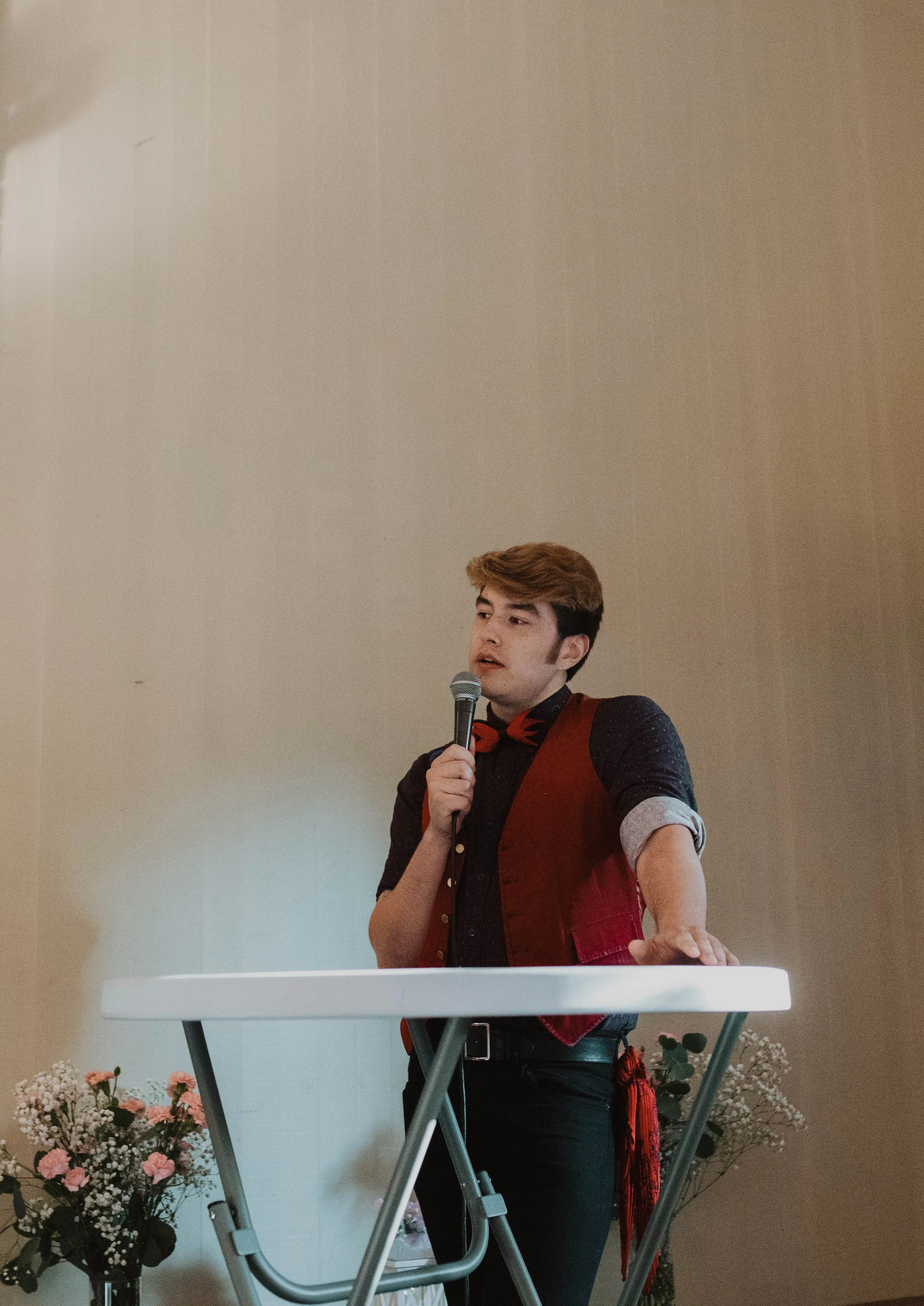 A young man with brown hair, dressed in a black shirt with rolled-up sleeves, a red vest, and a black belt, standing behind a white podium, holding a microphone, speaking to an audience. There are pink and white flowers on either side of the podium.