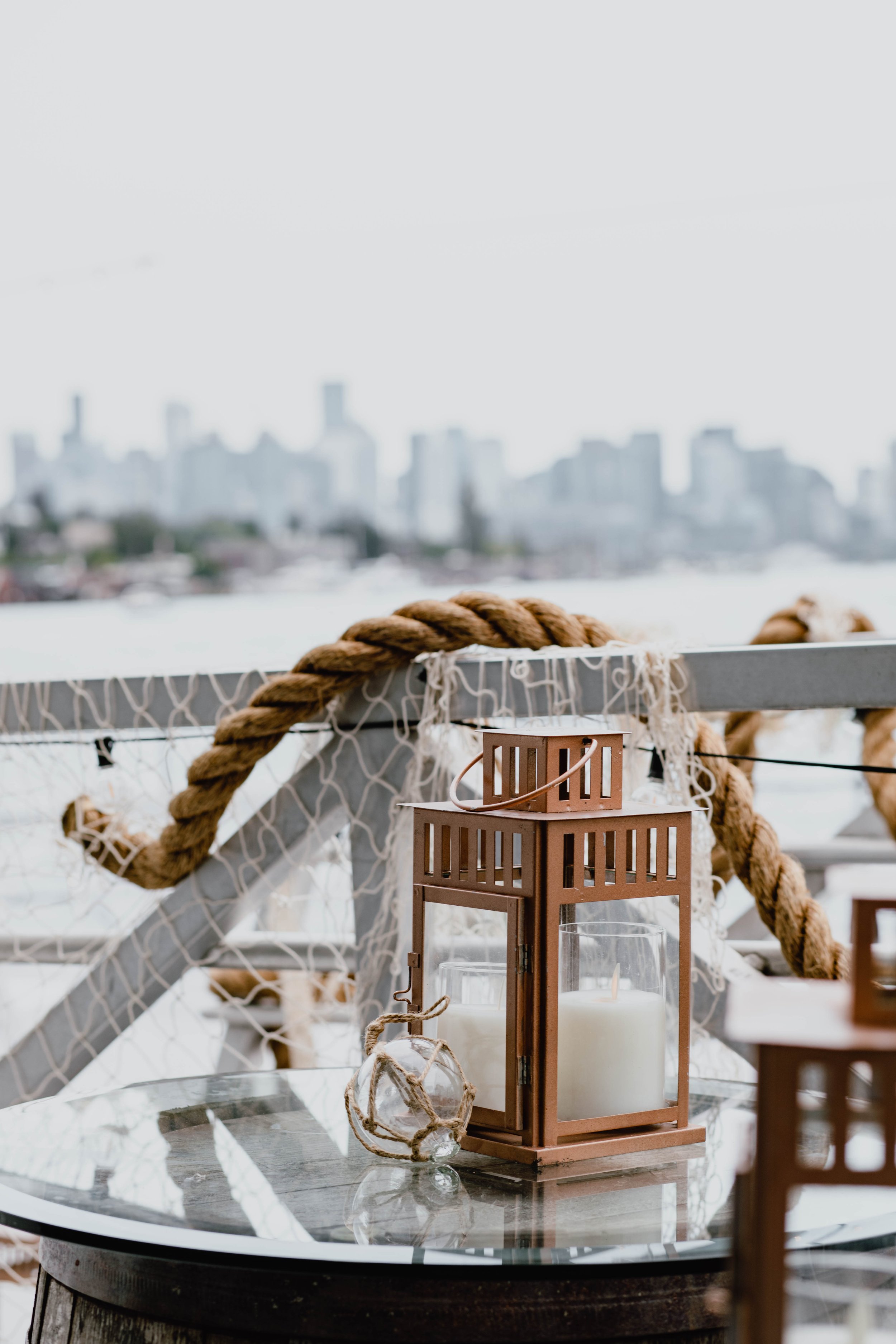Decorative candle lantern and a small glass sphere tied with jute twine on a glass-top table, with a nautical rope and netting in the background overlooking a city skyline. Seattle event photography