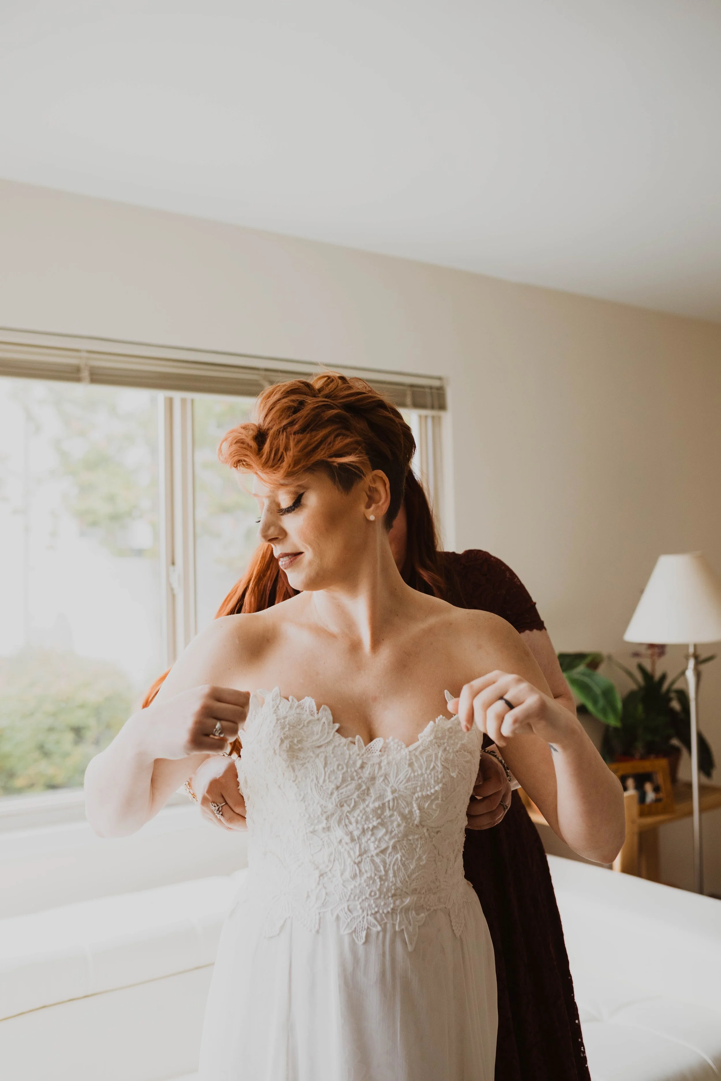 A bride getting ready, adjusting her white lace wedding dress with help from another person, in a bright room with large windows and a lamp in the background. Pioneer Square, Seattle, WA wedding photography.