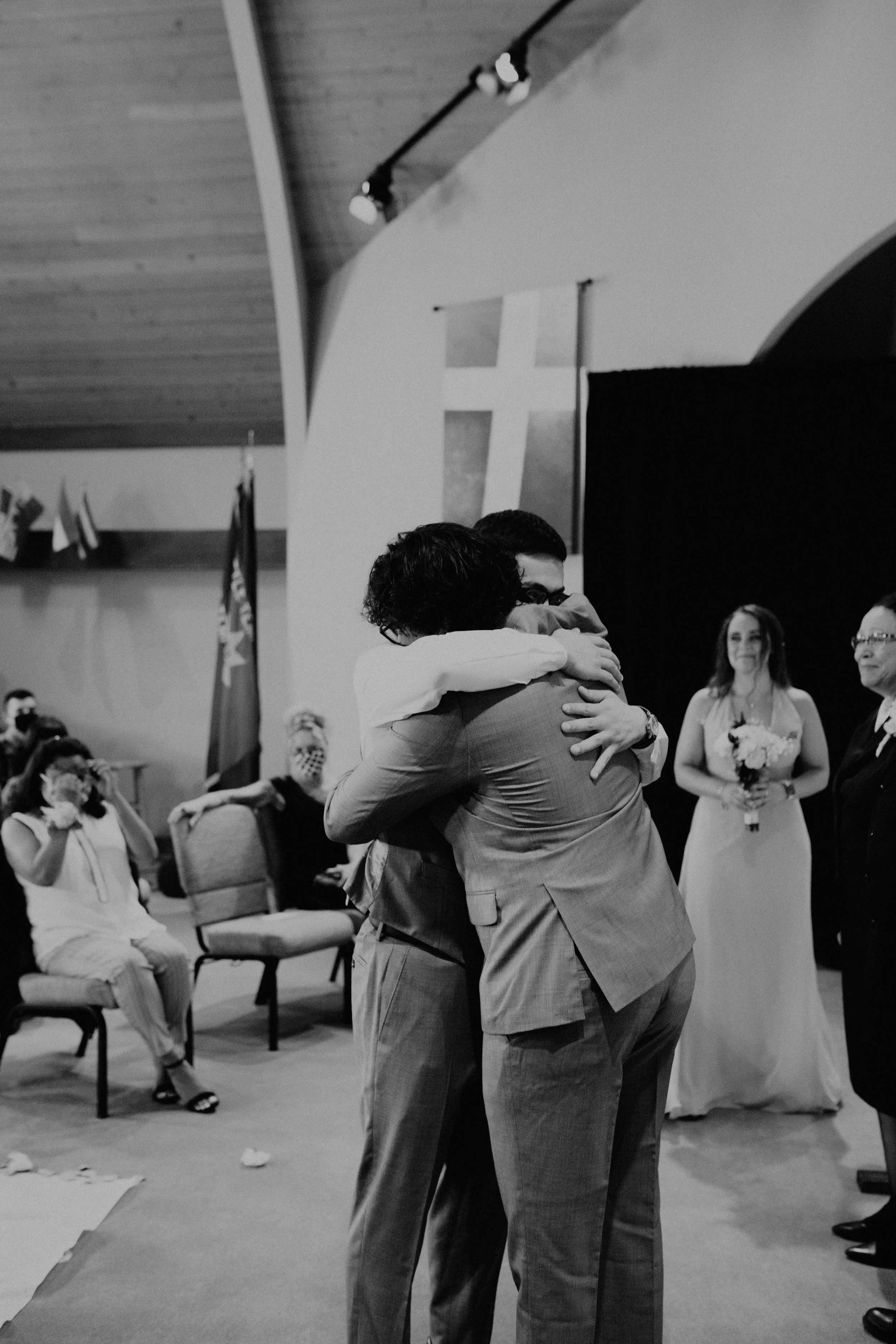 Two men in suits hugging each other at a wedding ceremony, with a woman in a wedding dress holding a bouquet and people taking photos in the background. Seattle, WA wedding photography.