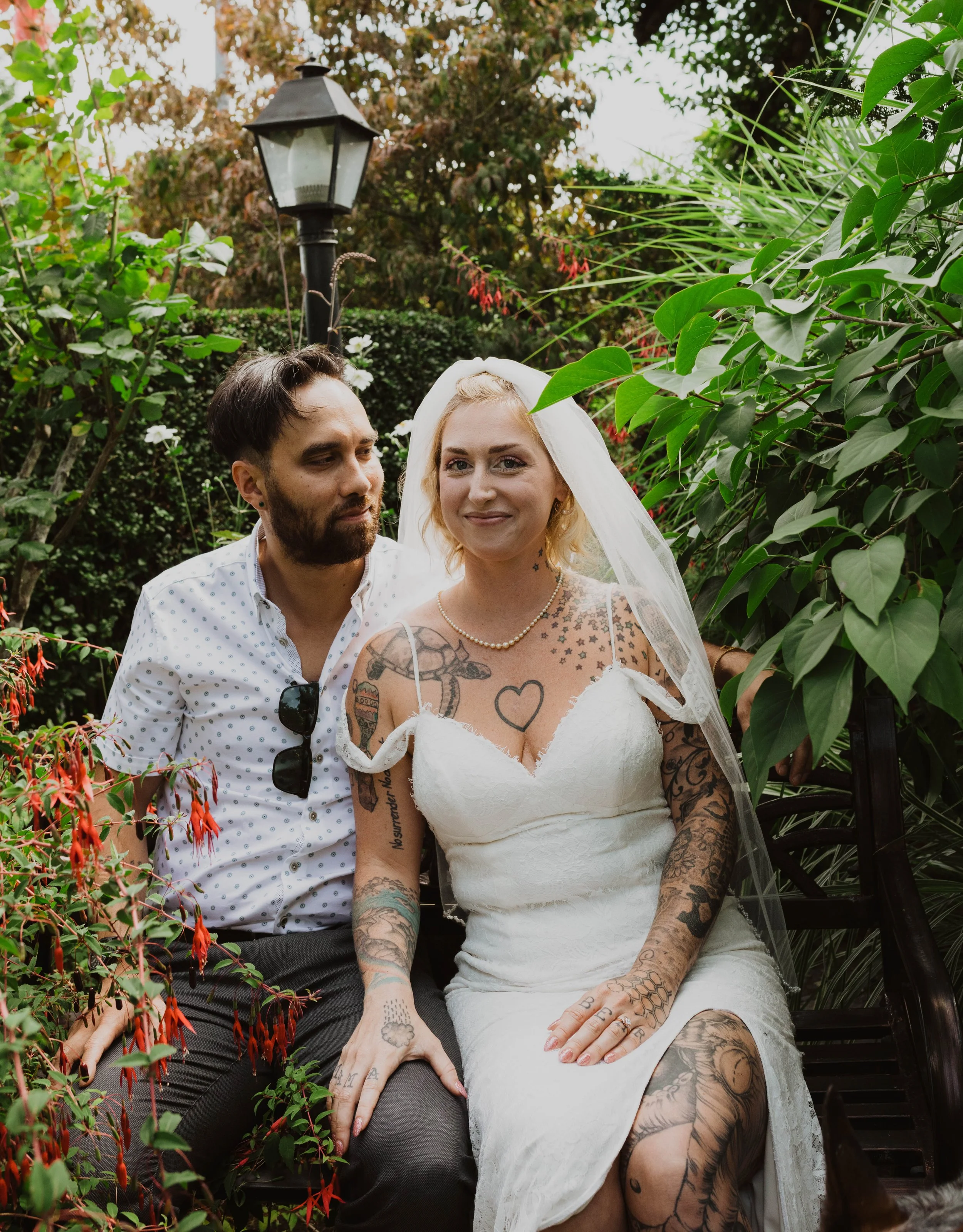 A wedding couple sitting on a park bench in a garden, the bride in a white dress and veil, the groom in a white shirt with sunglasses hanging from the chest pocket, surrounded by lush greenery and flowers. Seattle, WA wedding photography.