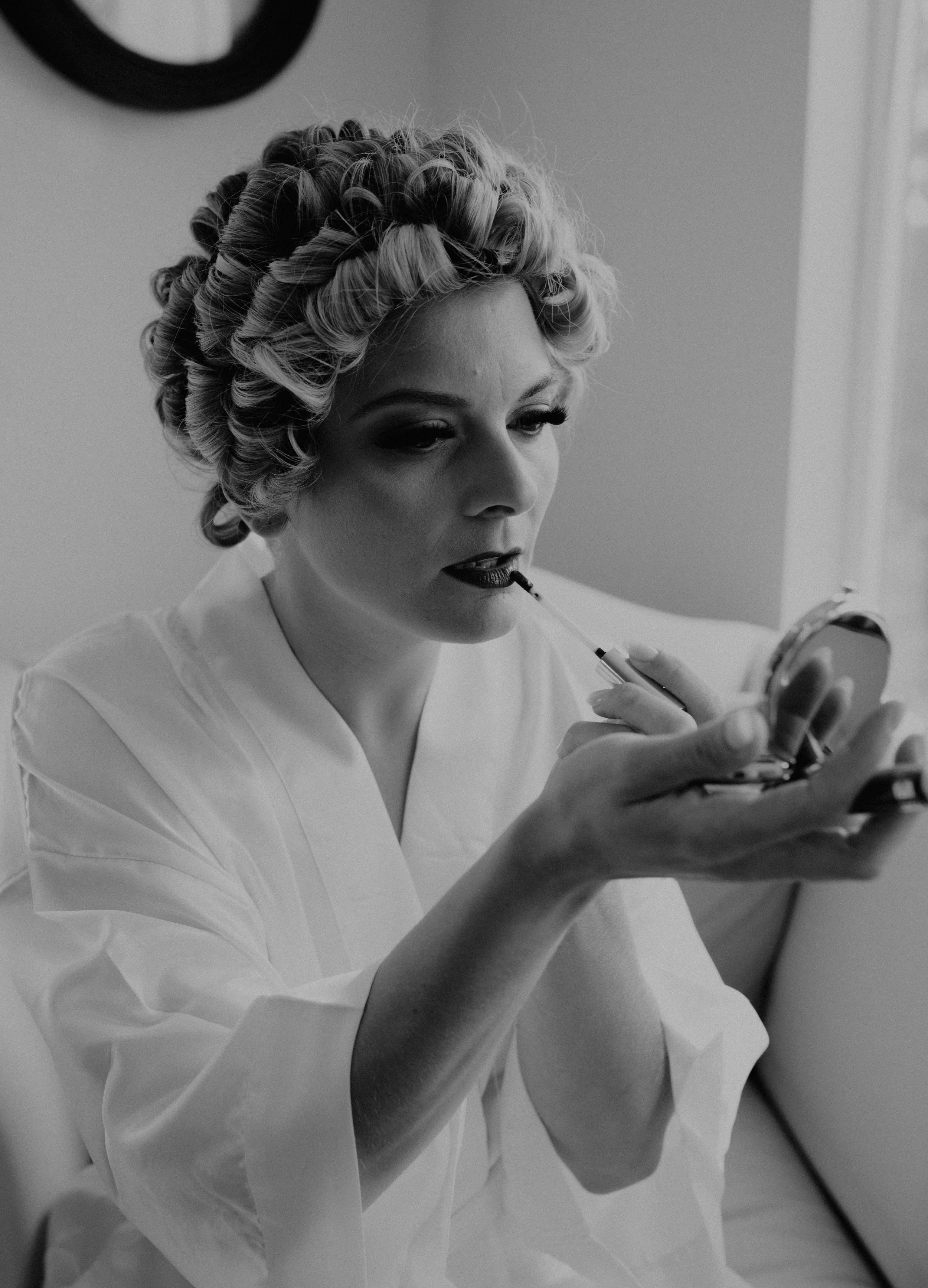 A woman with curled hair applying lipstick while checking her reflection in a mirror. Long Beach, WA wedding photography.