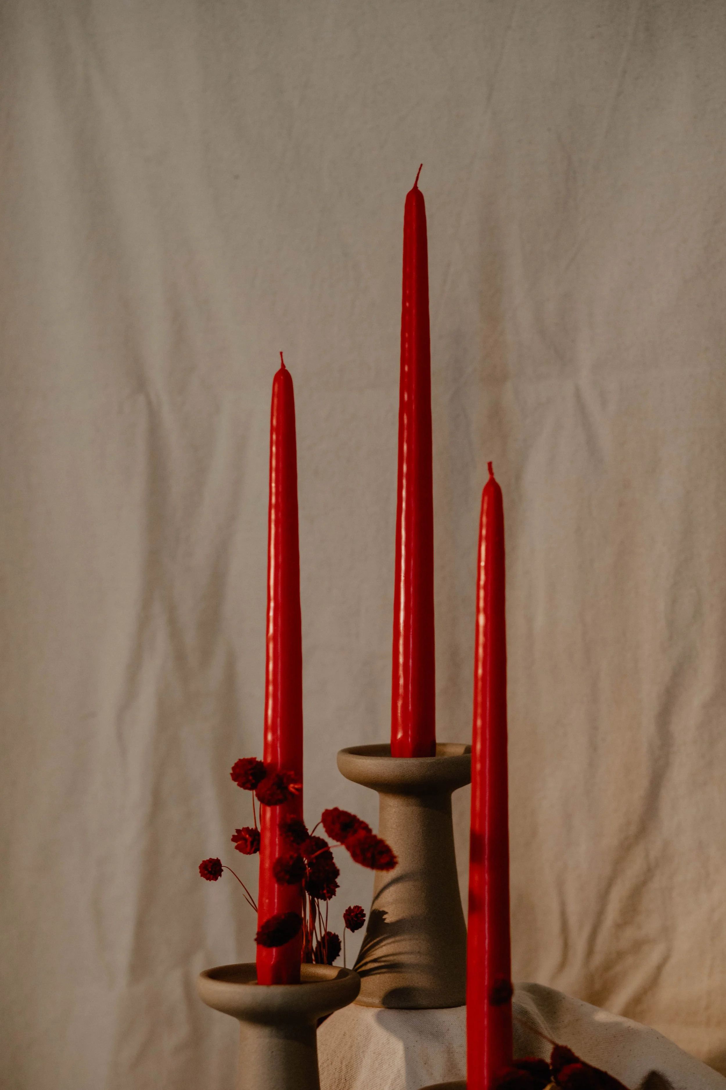 Three tall red candles on beige ceramic candleholders with dried red flowers in front, against a beige textured wall.  Seattle product photography