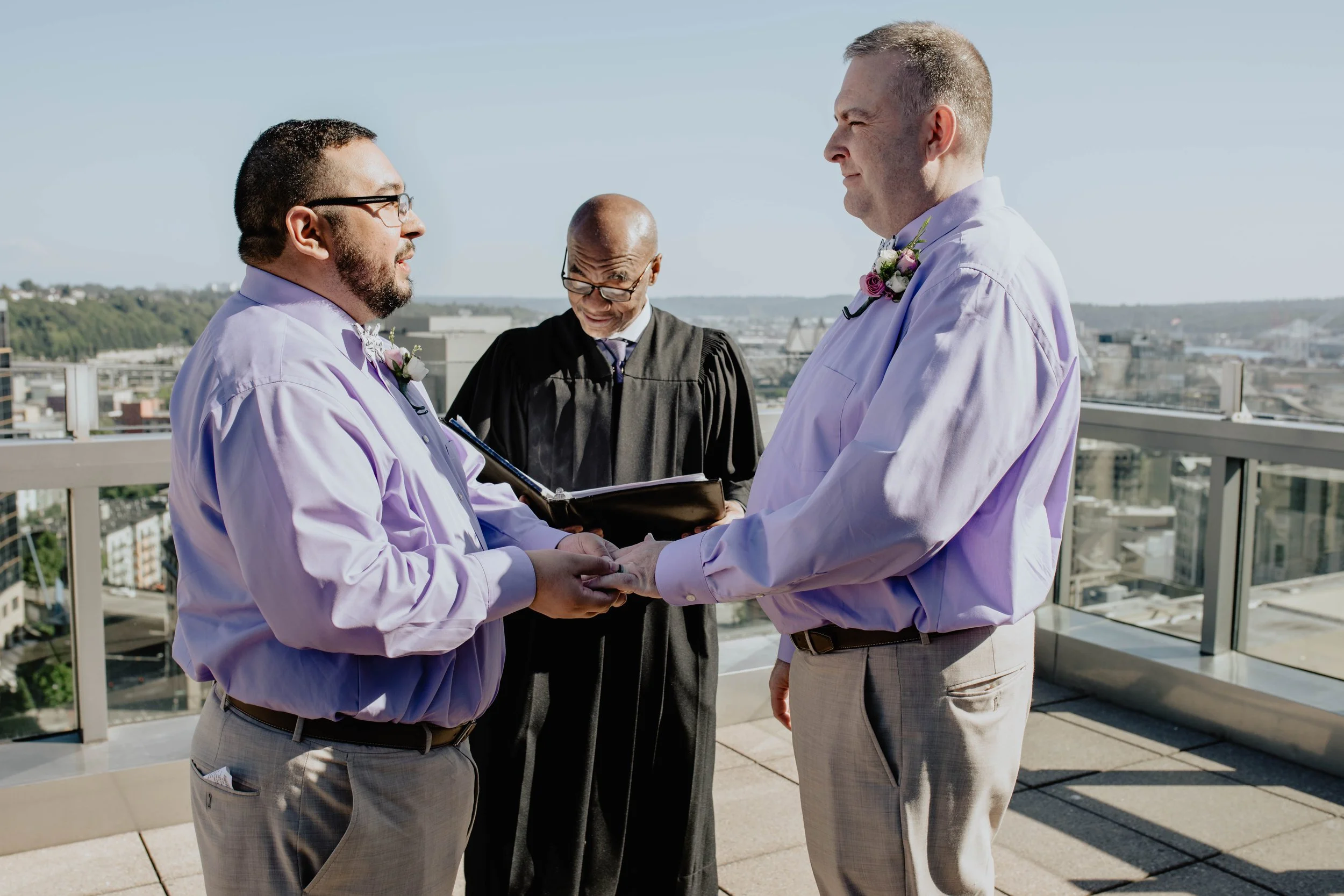 Two men in light purple shirts holding hands during a wedding ceremony on a rooftop, with a officiant in black robes reading from a book, and a city skyline in the background. Seattle Municipal Courthouse wedding photography.