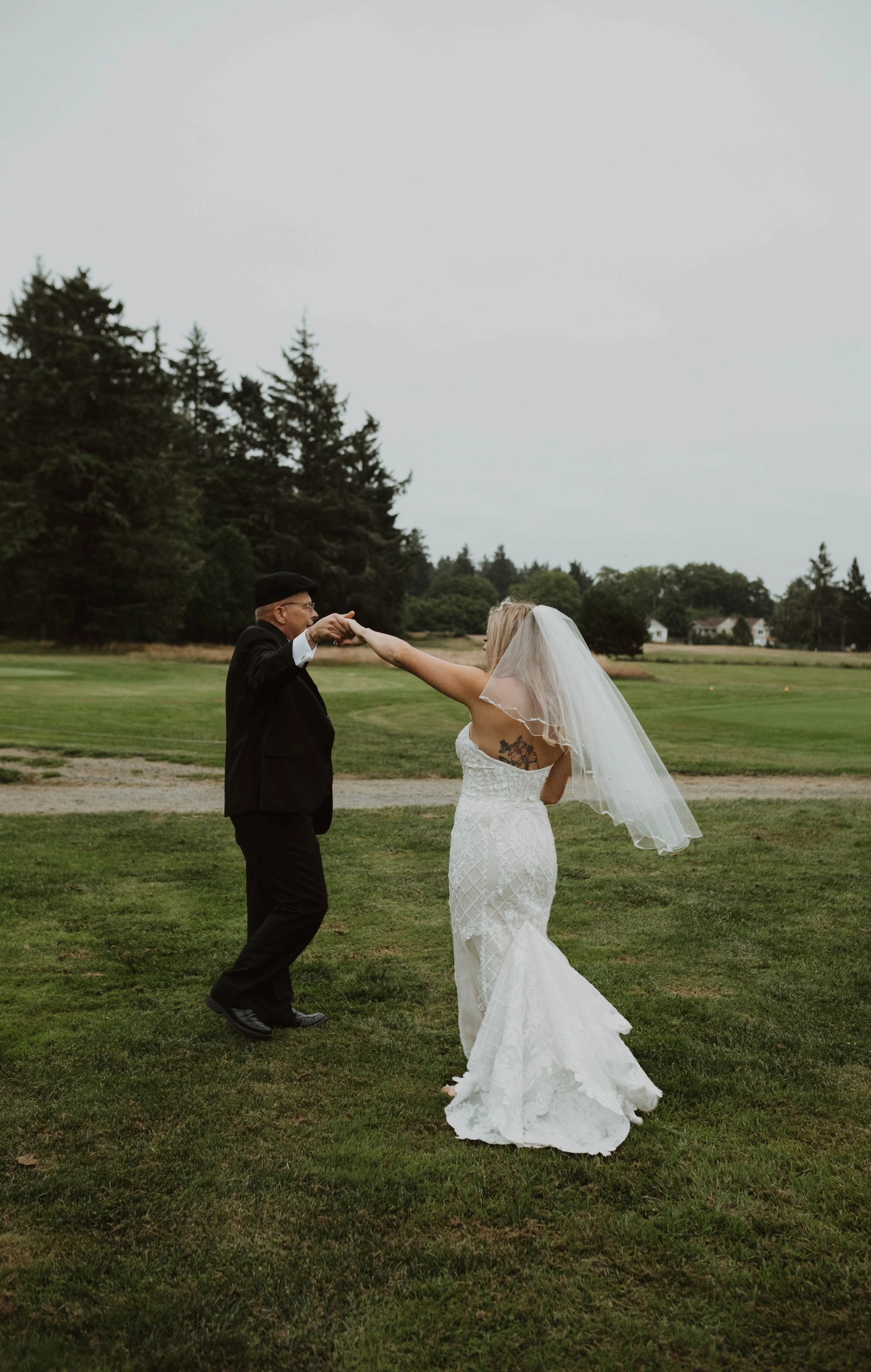 A bride in a white lace wedding dress and veil dancing with an elderly man in a black suit and hat on a grassy field outdoors with trees and houses in the background. Long Beach, WA wedding photography.