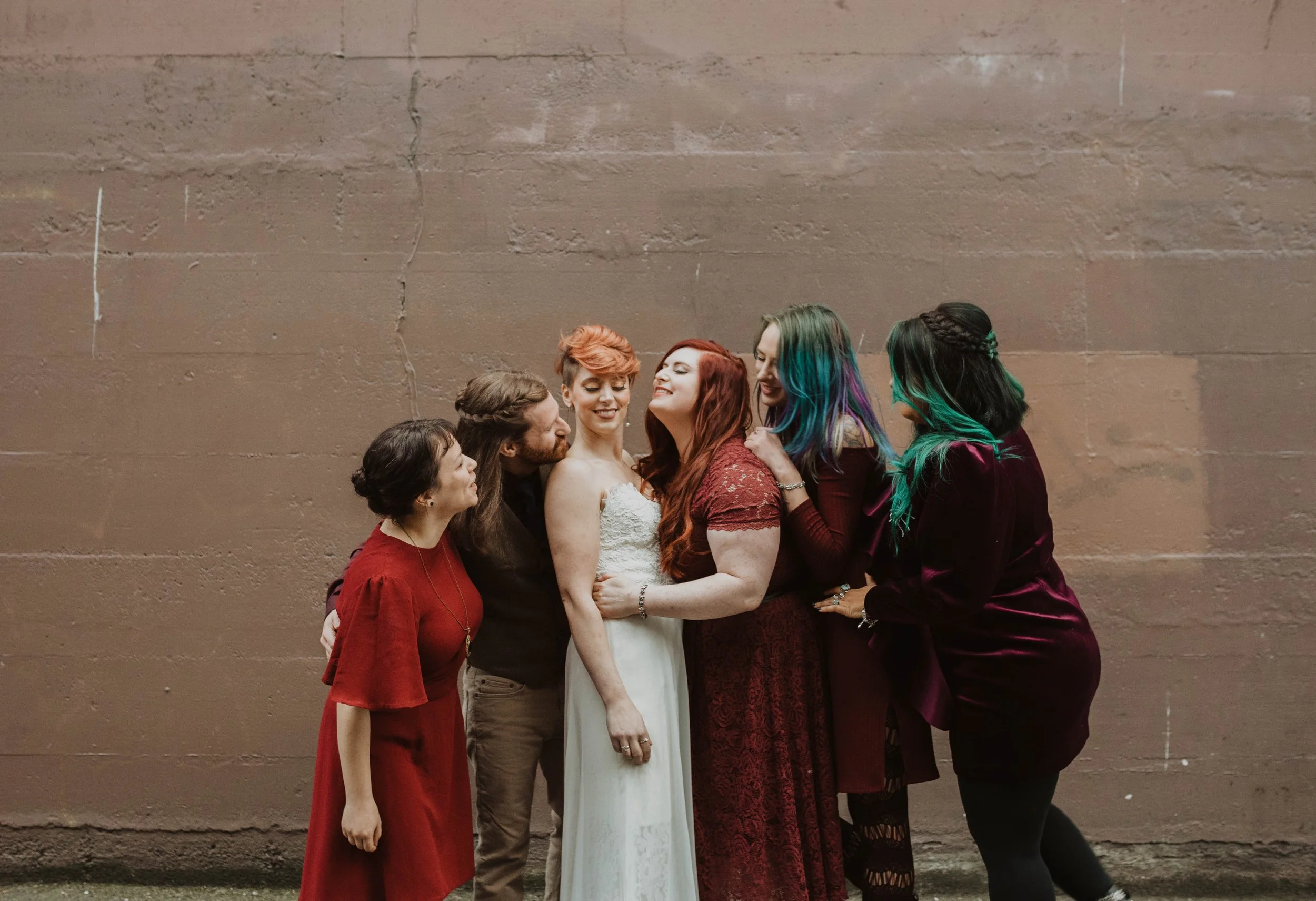 Group of six people, including a bride in a white dress, standing close and smiling against a large brown wall. Pioneer Square, Seattle, WA wedding photography.