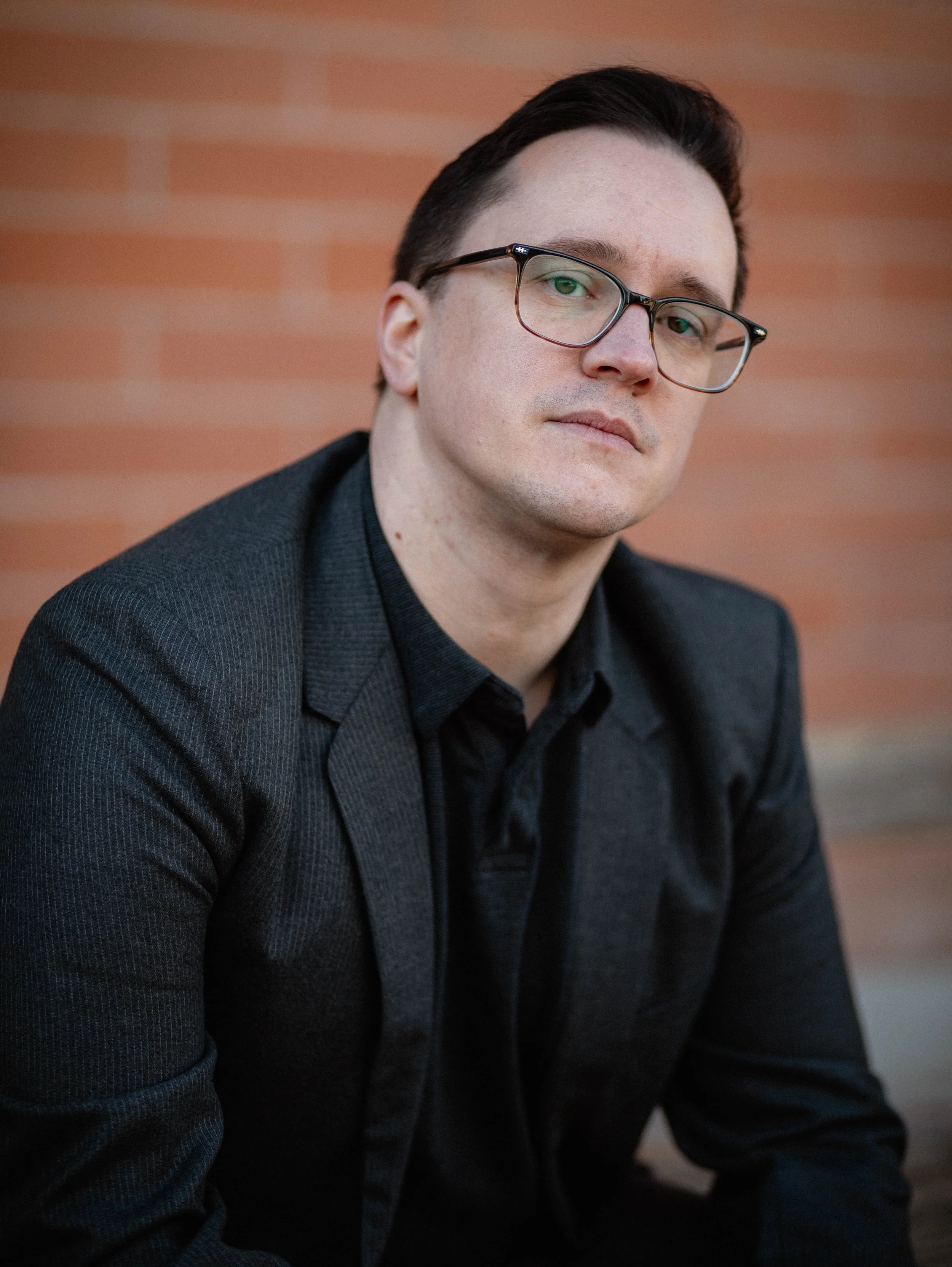 A young man with dark hair, wearing glasses and a dark suit, sitting outdoors against a brick wall, looking at the camera. Seattle professional head shot photography