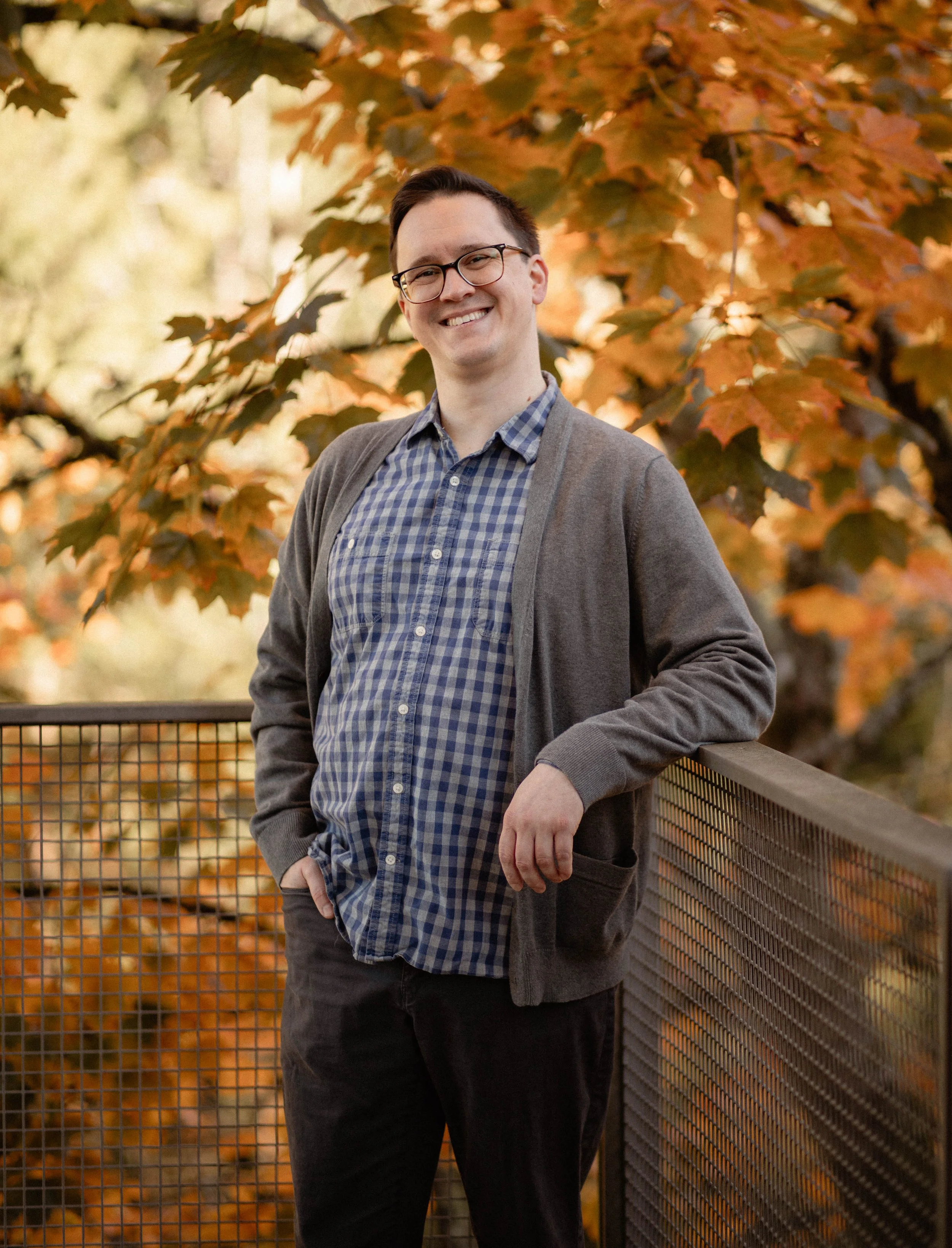 A young man with glasses and short dark hair, smiling, standing outdoors on a bridge with autumn leaves in the background. He is wearing a checkered shirt and a gray cardigan. Seattle professional head shot photography
