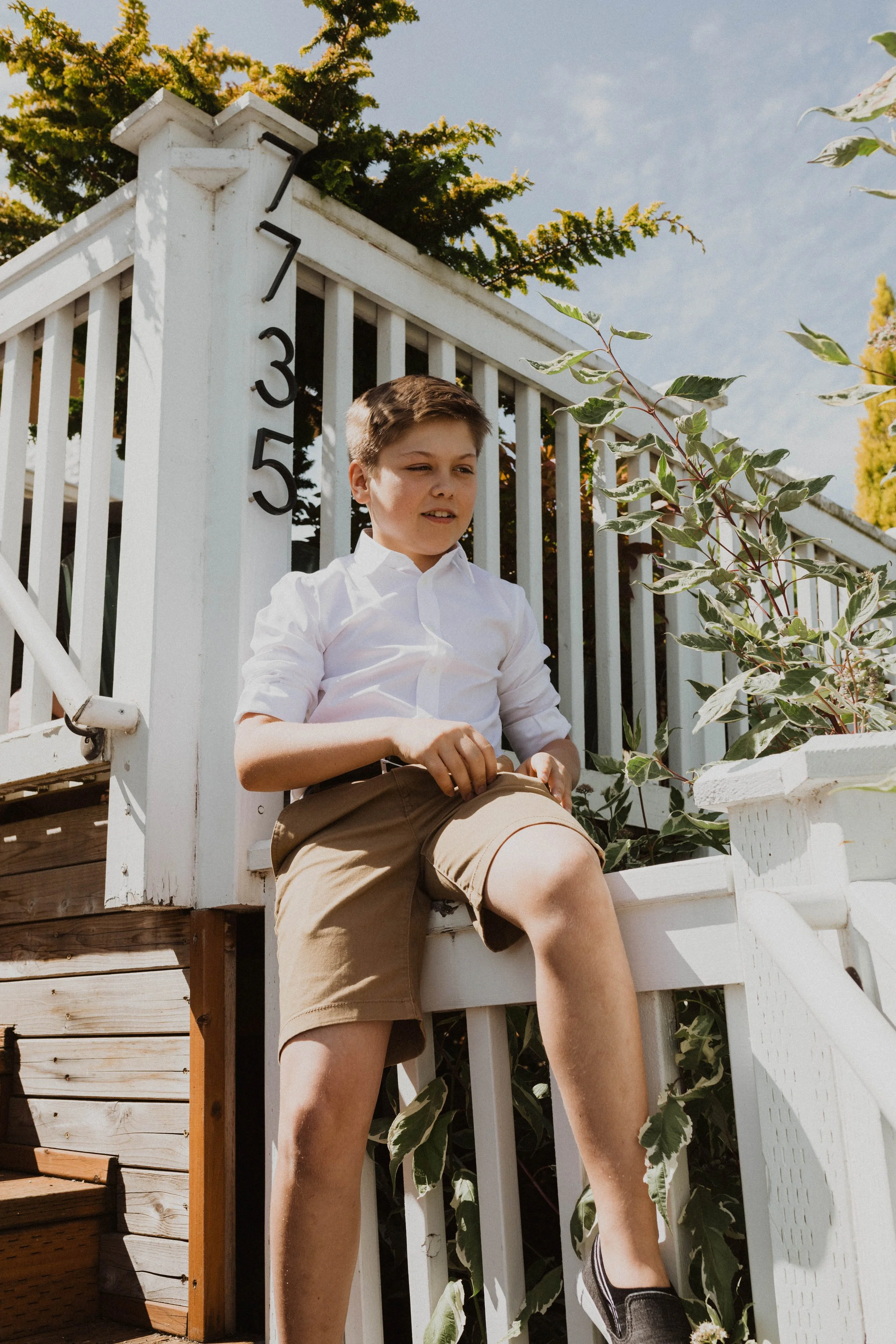 A young boy sitting on a staircase outside near a garden with greenery. He is wearing a white shirt and khaki shorts, with one leg hanging down and the other bent. The house number 735 is visible on a post near him. Seattle, WA wedding photography.