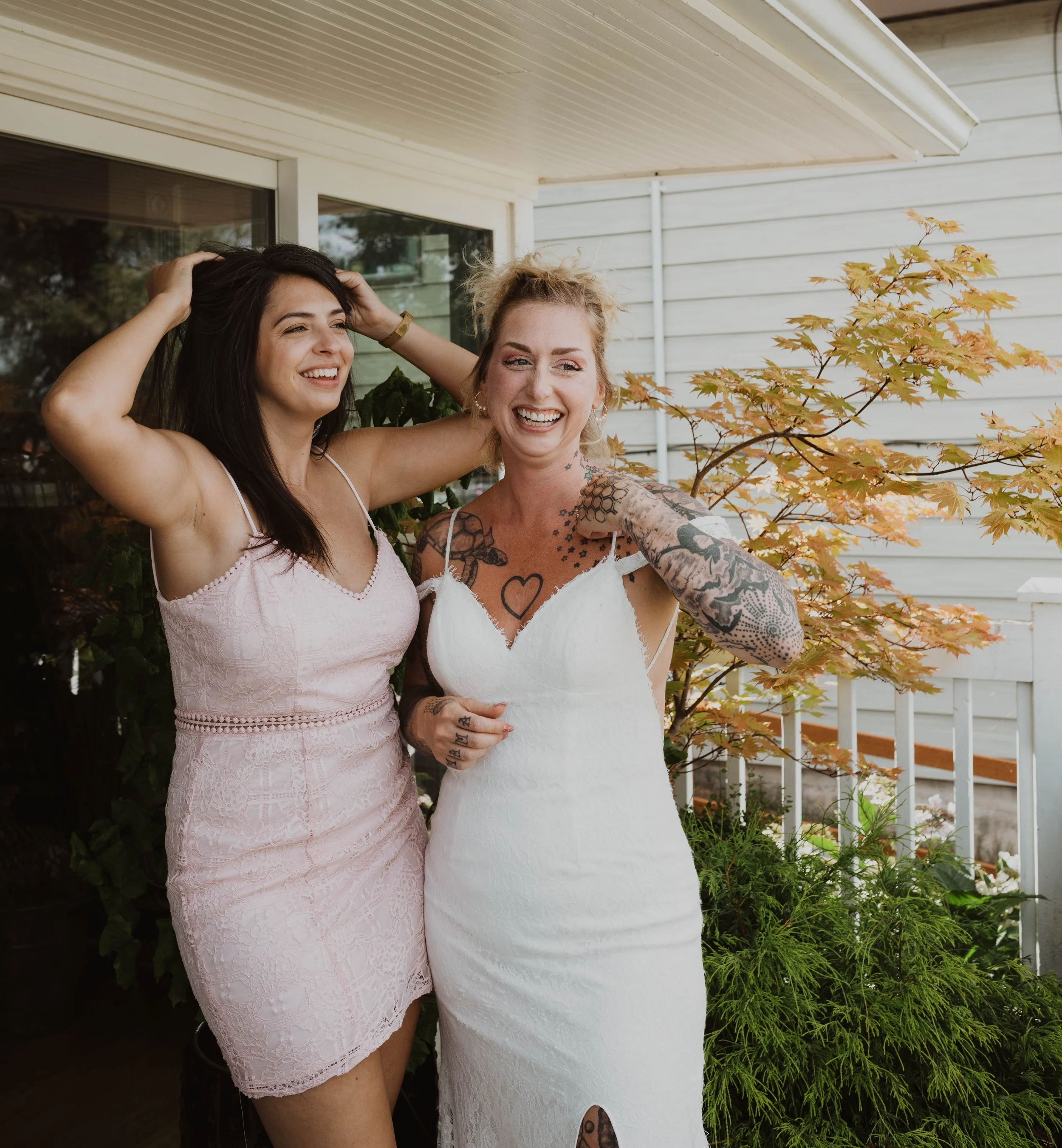 Two women smiling and laughing outdoors on a porch with plants and trees, one wearing a pink dress and the other in a white dress with tattoos. Seattle, WA wedding photography.