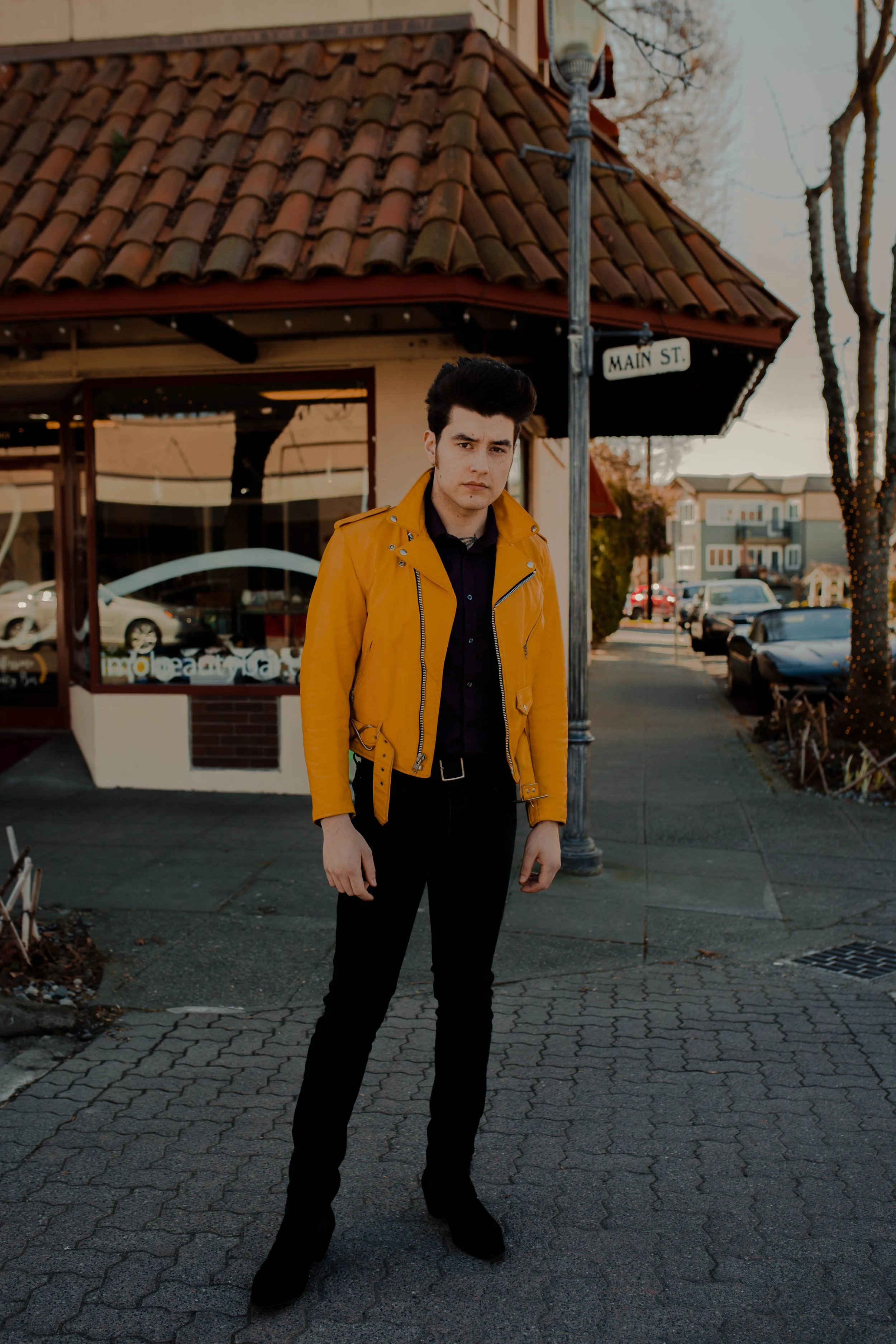 Young man standing on sidewalk in front of building with tiled roof, wearing yellow jacket and black pants, with street sign reading 'Main St' nearby. Seattle professional head shot photography
