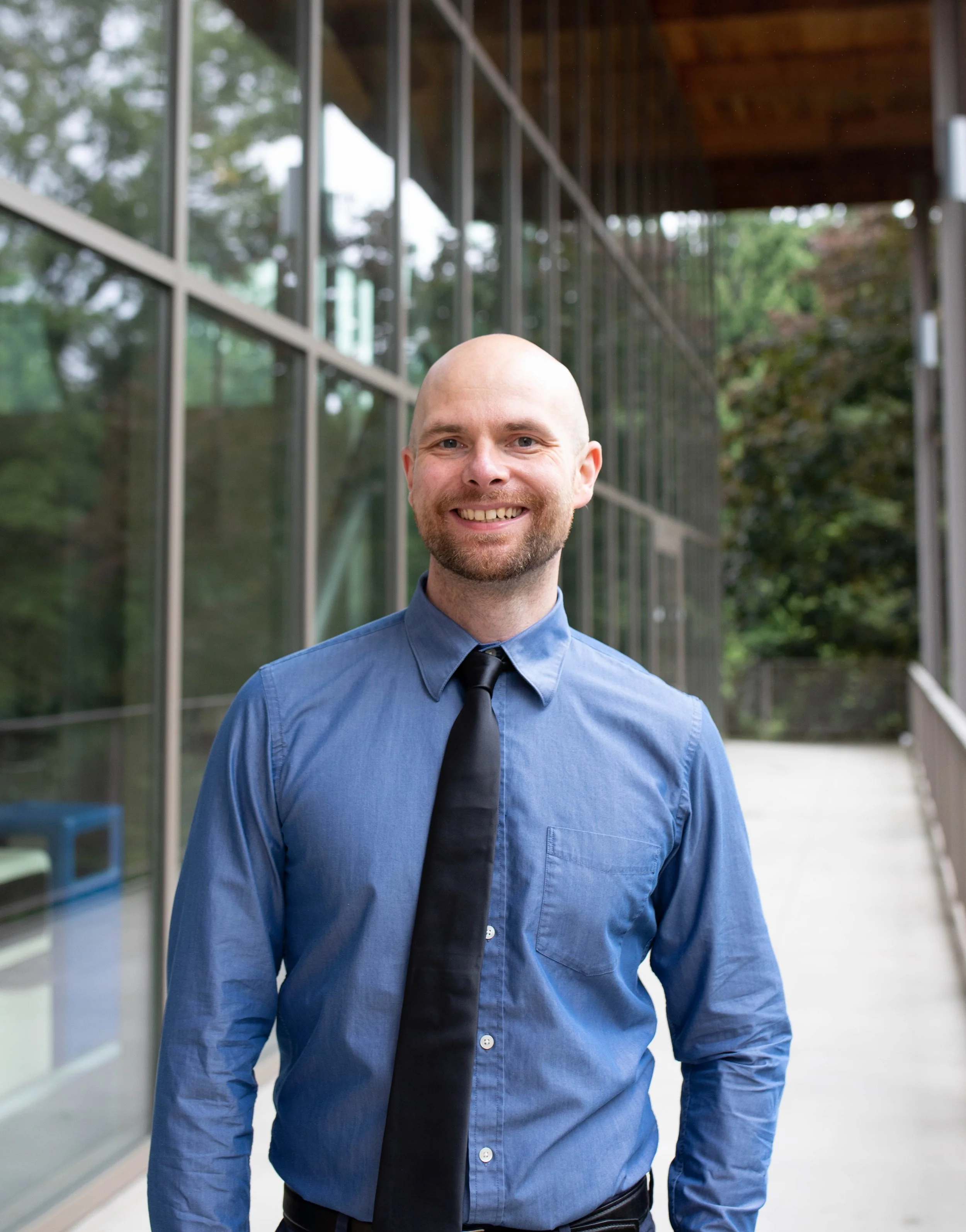 A smiling, bearded man in a blue dress shirt and black tie standing outside near a modern glass building with trees in the background. Seattle professional head shot photography