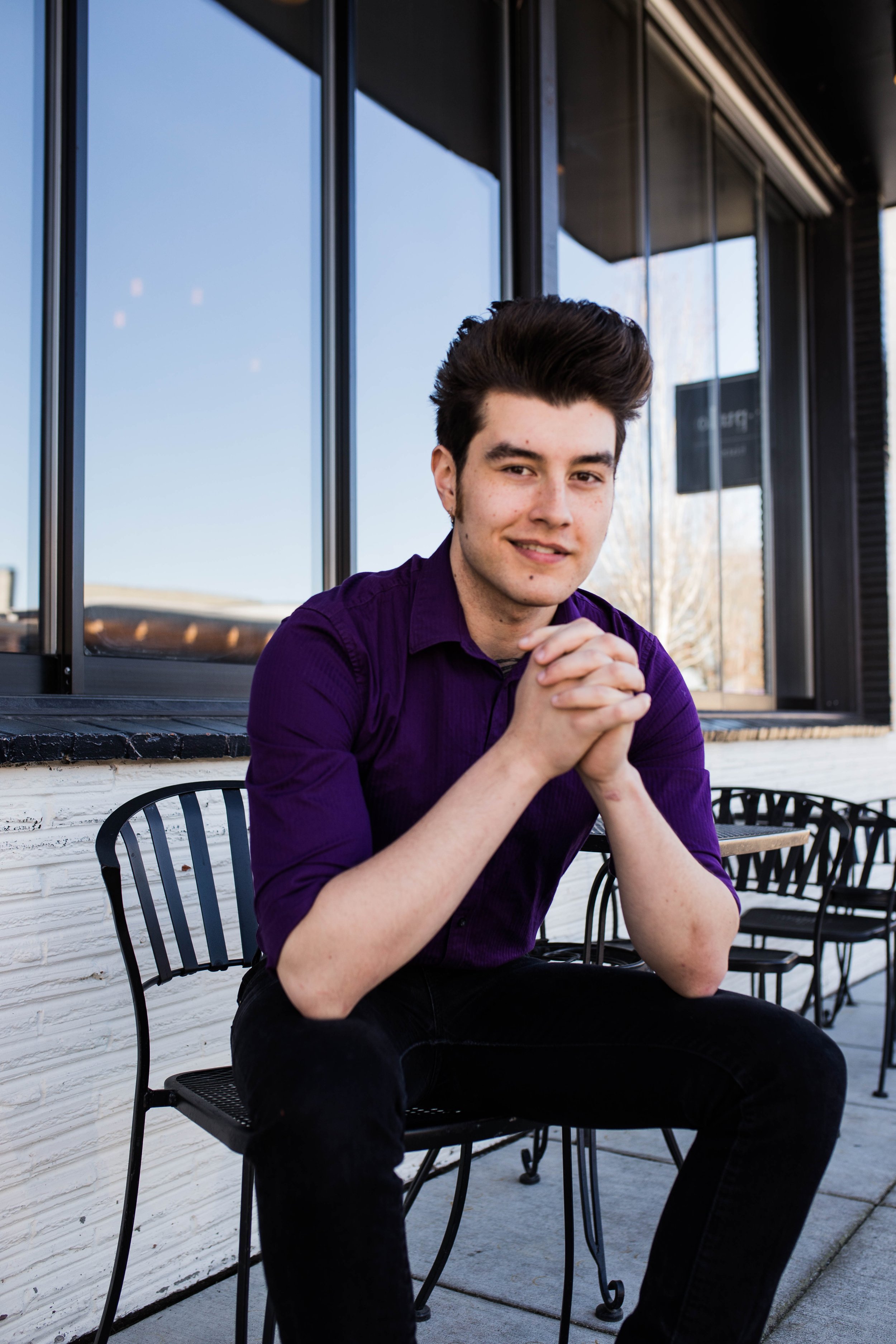A young man with dark hair, wearing a purple shirt, sitting outside in front of a building with large glass windows, smiling and clasping his hands. Seattle professional head shot photography