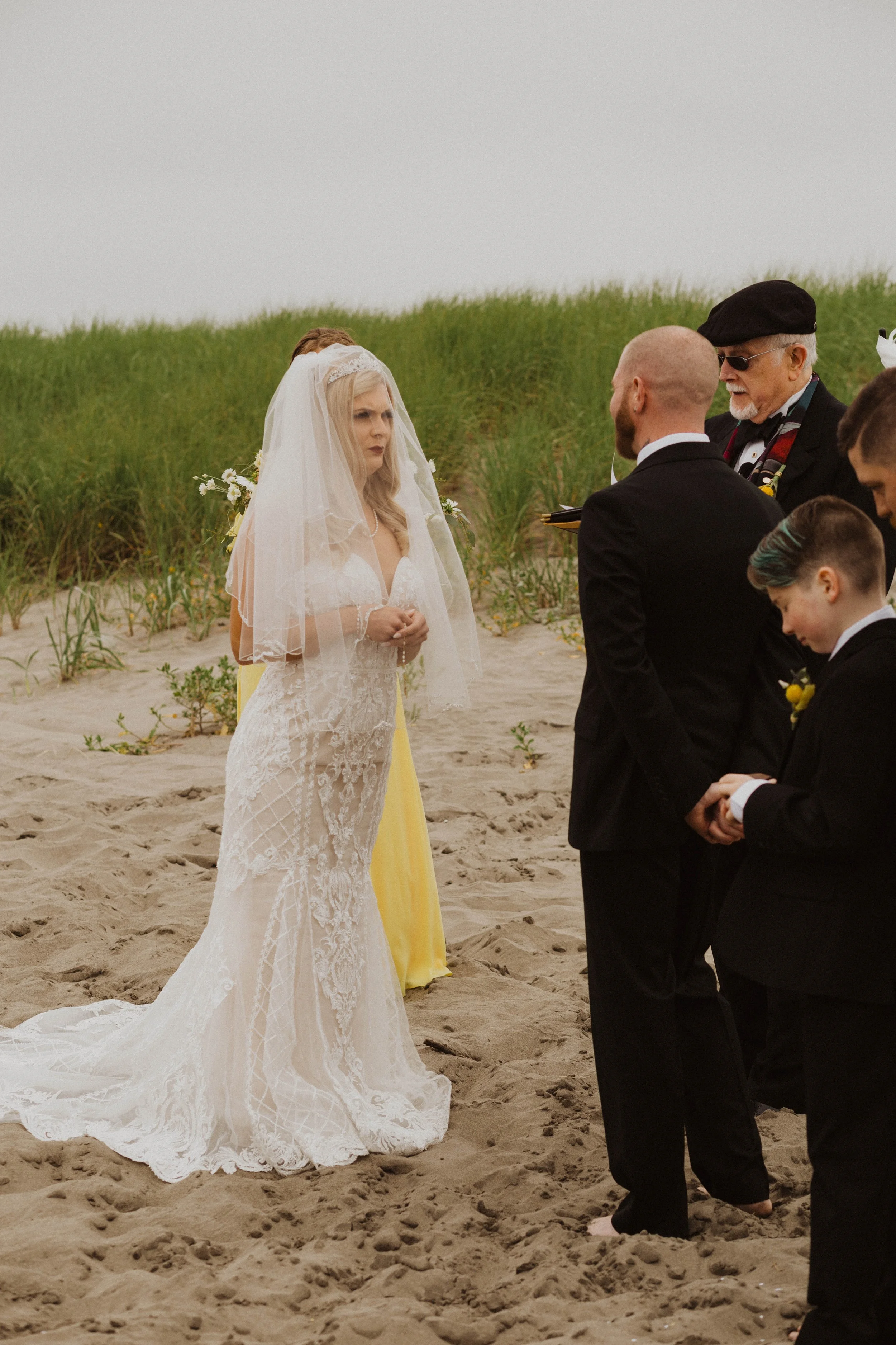 A bride with long blonde hair, wearing a lace wedding dress and a veil, faces a groom and officiant during a beach wedding ceremony, with grass and sand in the background. Long Beach, WA wedding photography.