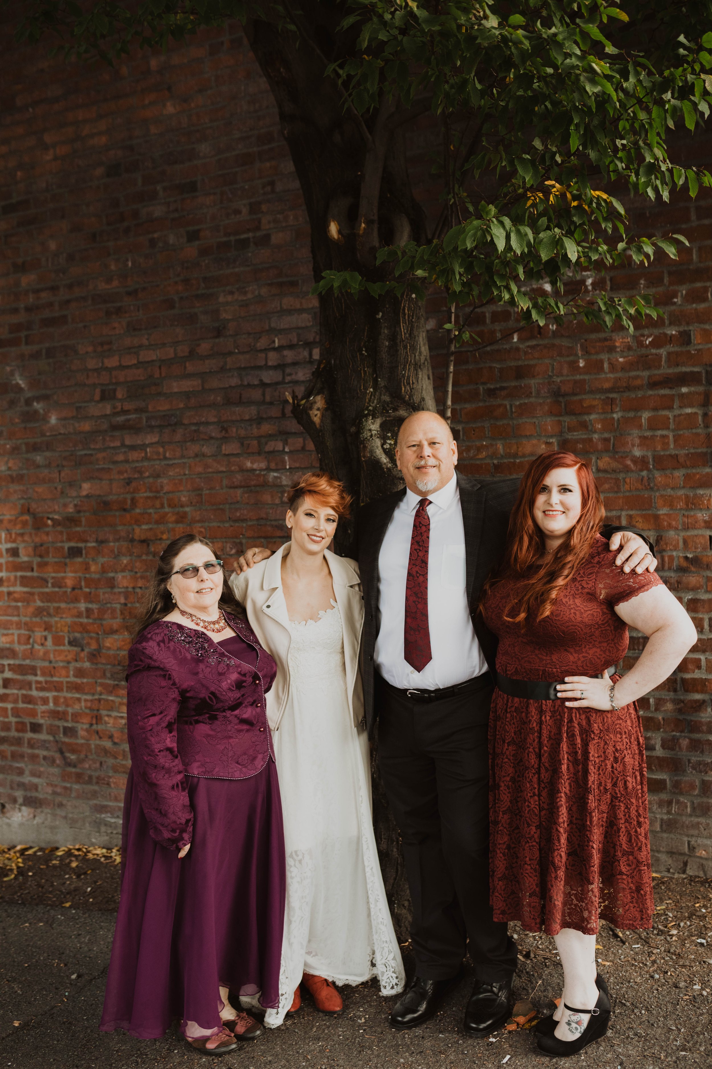 Four people standing outdoors beside a tree, smiling, with a brick wall in the background. They are dressed in formal and semi-formal attire. Pioneer Square, Seattle, WA wedding photography.