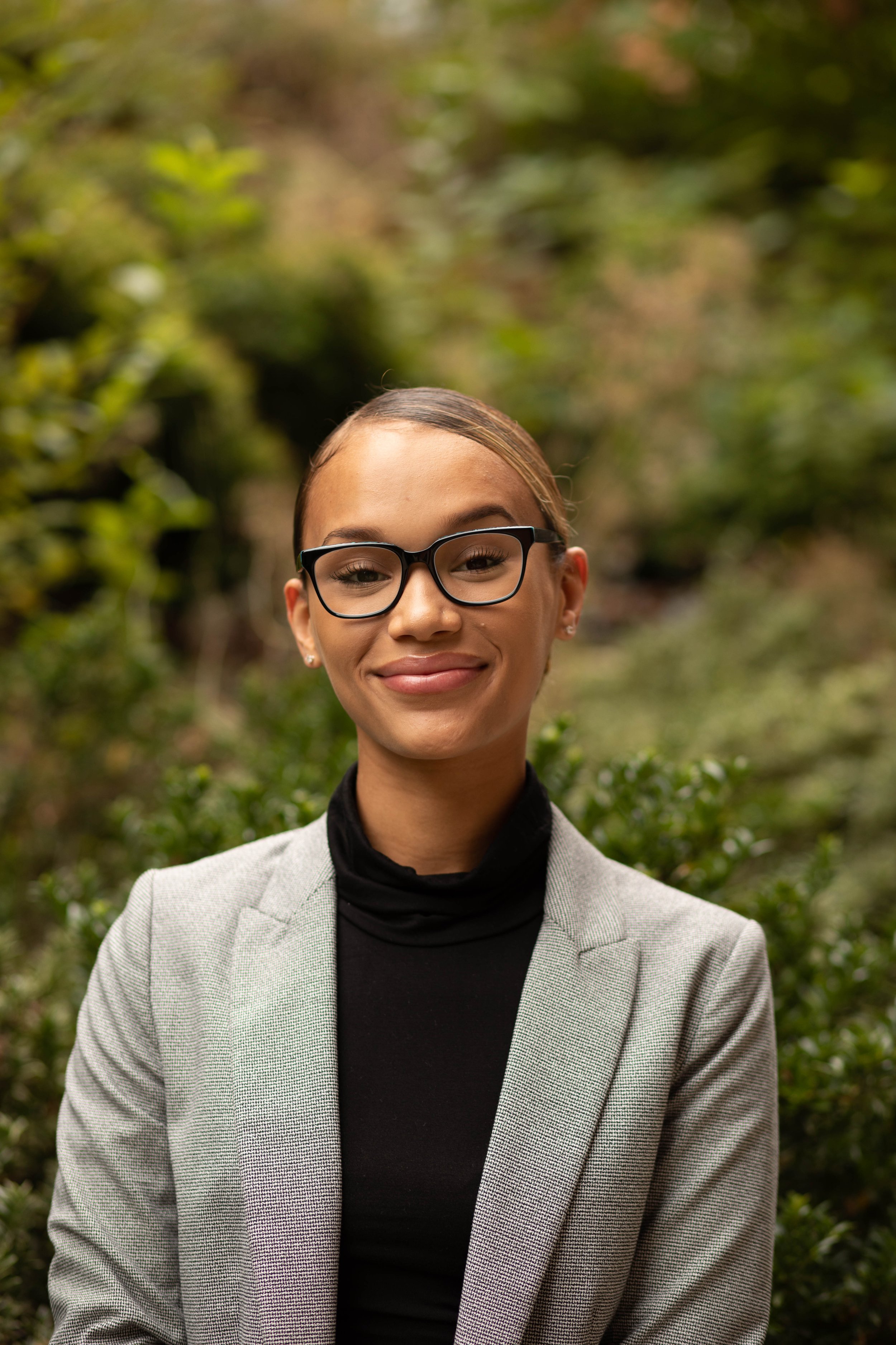 A young woman with glasses, wearing a light gray blazer and black turtleneck, smiling outdoors in front of green foliage. Seattle professional head shot photography