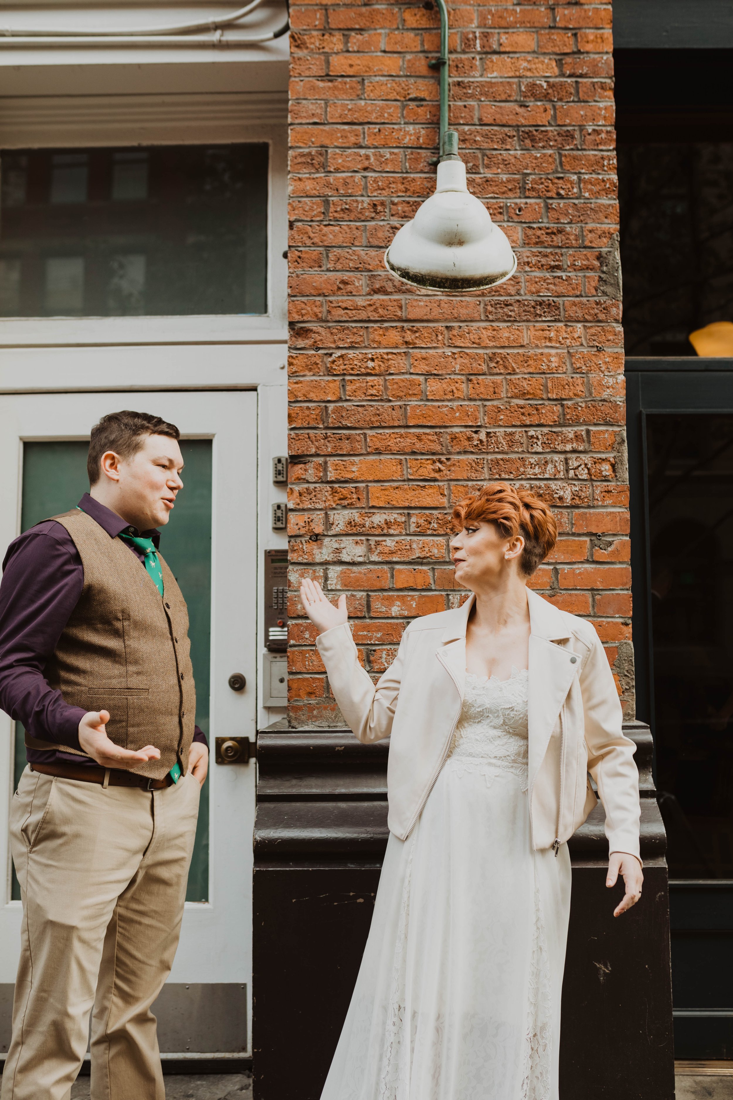 Two people standing outside a brick building, engaging in a conversation. One is a man in a vest and khakis, and the other is a woman in a white dress and a light-colored jacket. Pioneer Square, Seattle, WA wedding photography.