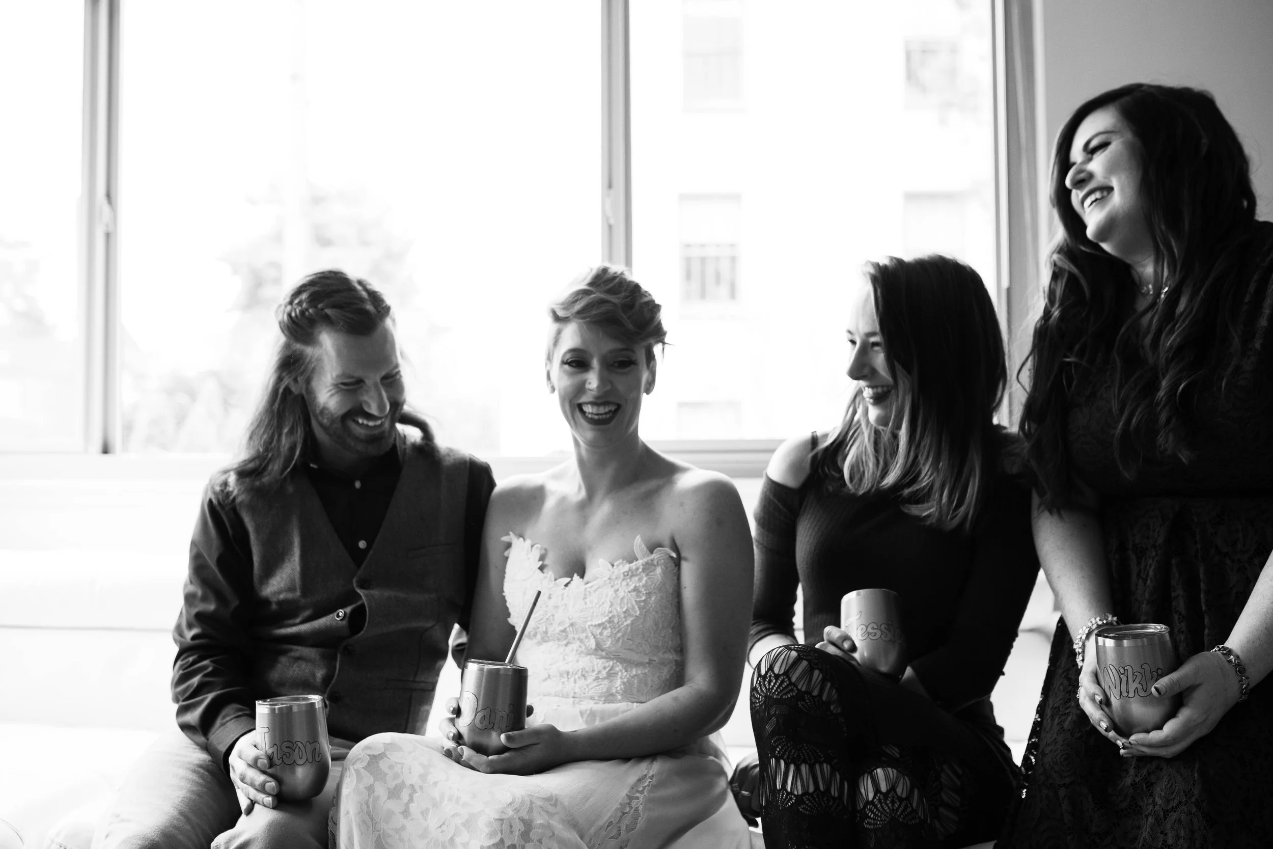 Four women sitting together indoors, laughing and enjoying drinks, with large windows in the background. Pioneer Square, Seattle, WA wedding photography.