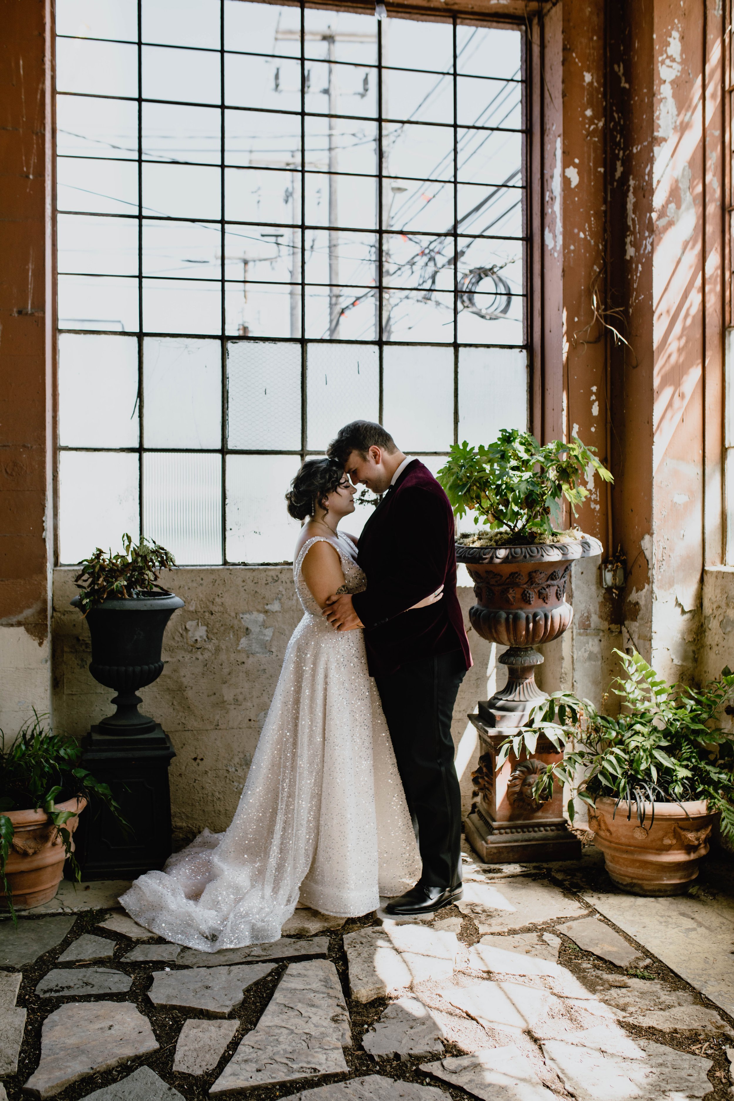 A couple in wedding attire stands close together inside an industrial-style building with large windows and potted plants, sharing an intimate moment. Wedding photography at The Ruins venue, in Queen Anne Seattle, WA
