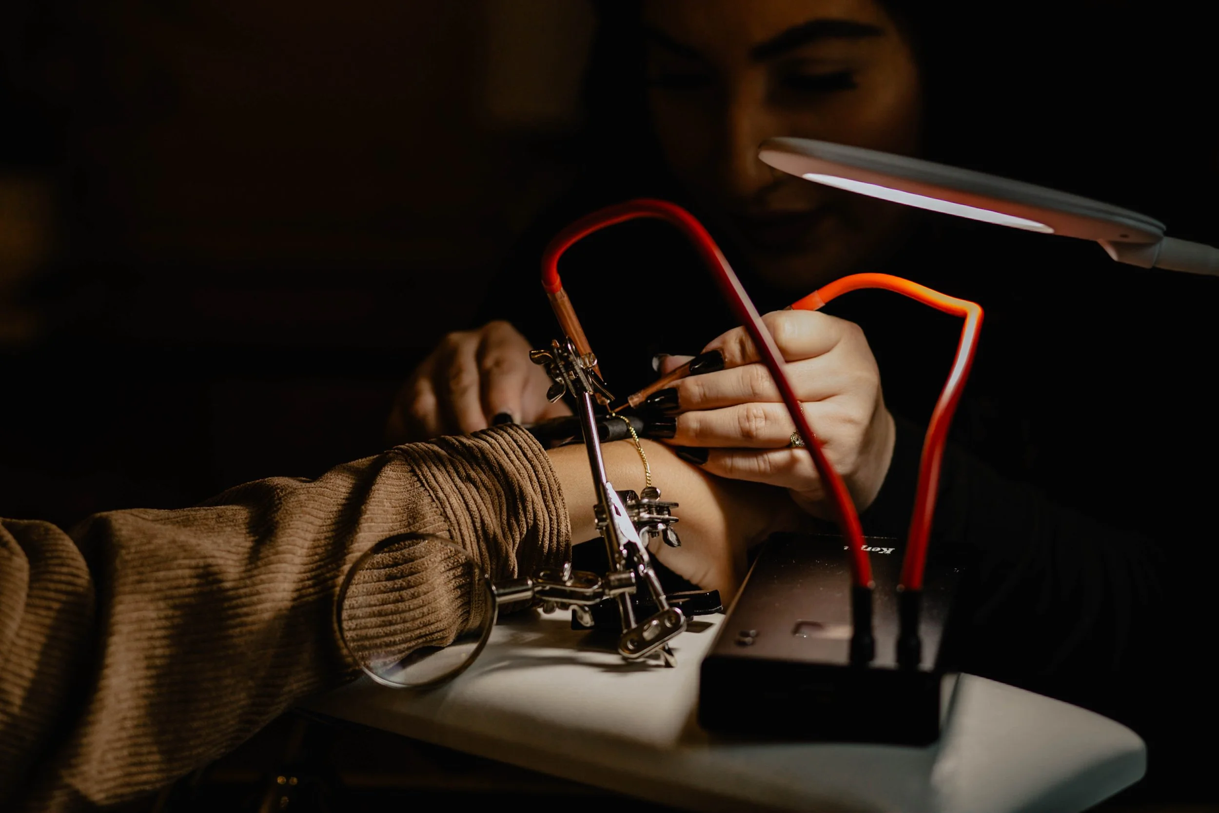 A person with long dark hair and multiple rings on fingers works on a person's arm using a device, with a lamp providing light, while an arm tattoo is visible on the arm being worked on, in a dimly lit setting. Seattle professional head shot photogra