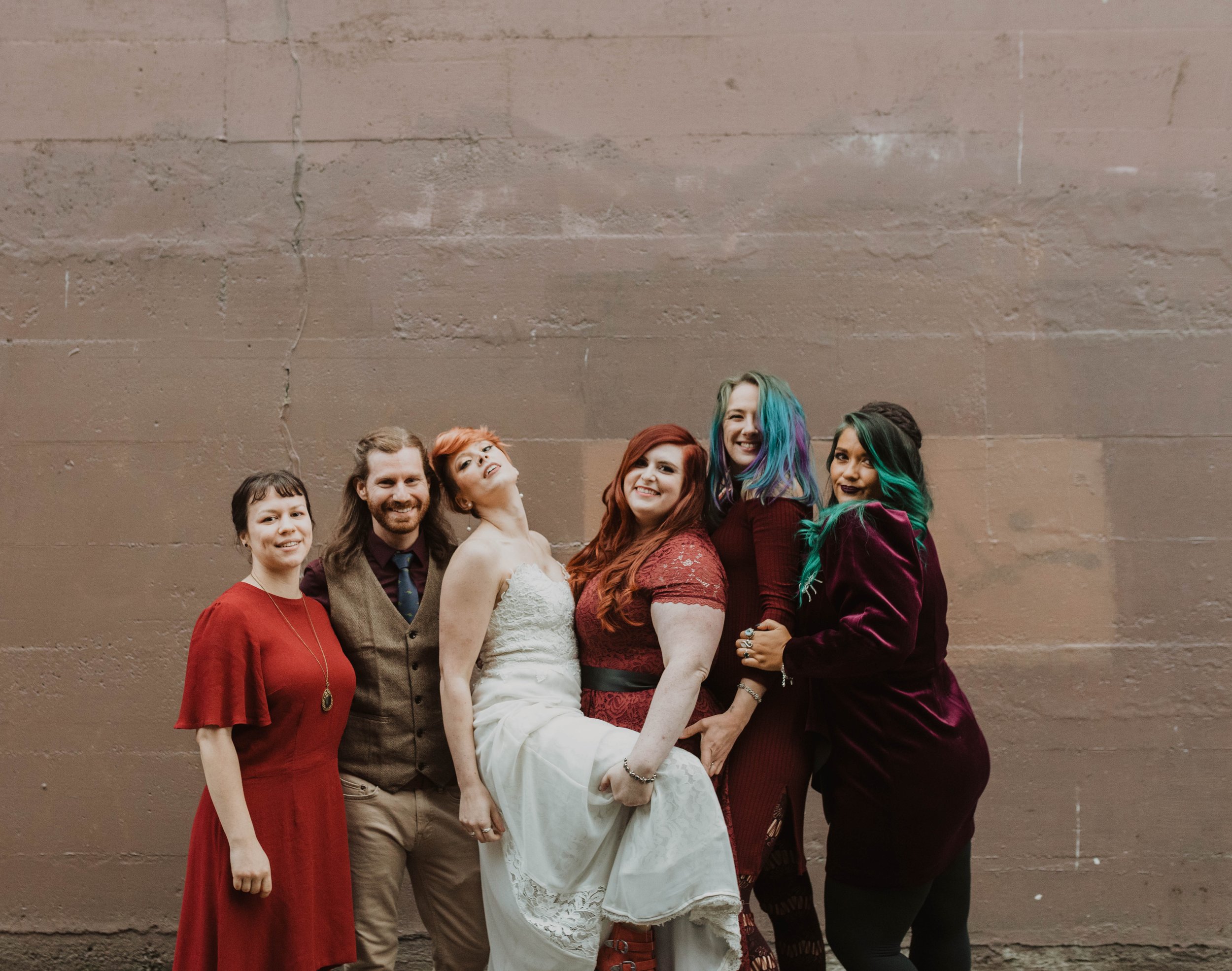 Group of six diverse people smiling and posing against a plain brown wall, with the woman in the white wedding dress in the center. Pioneer Square, Seattle, WA wedding photography.