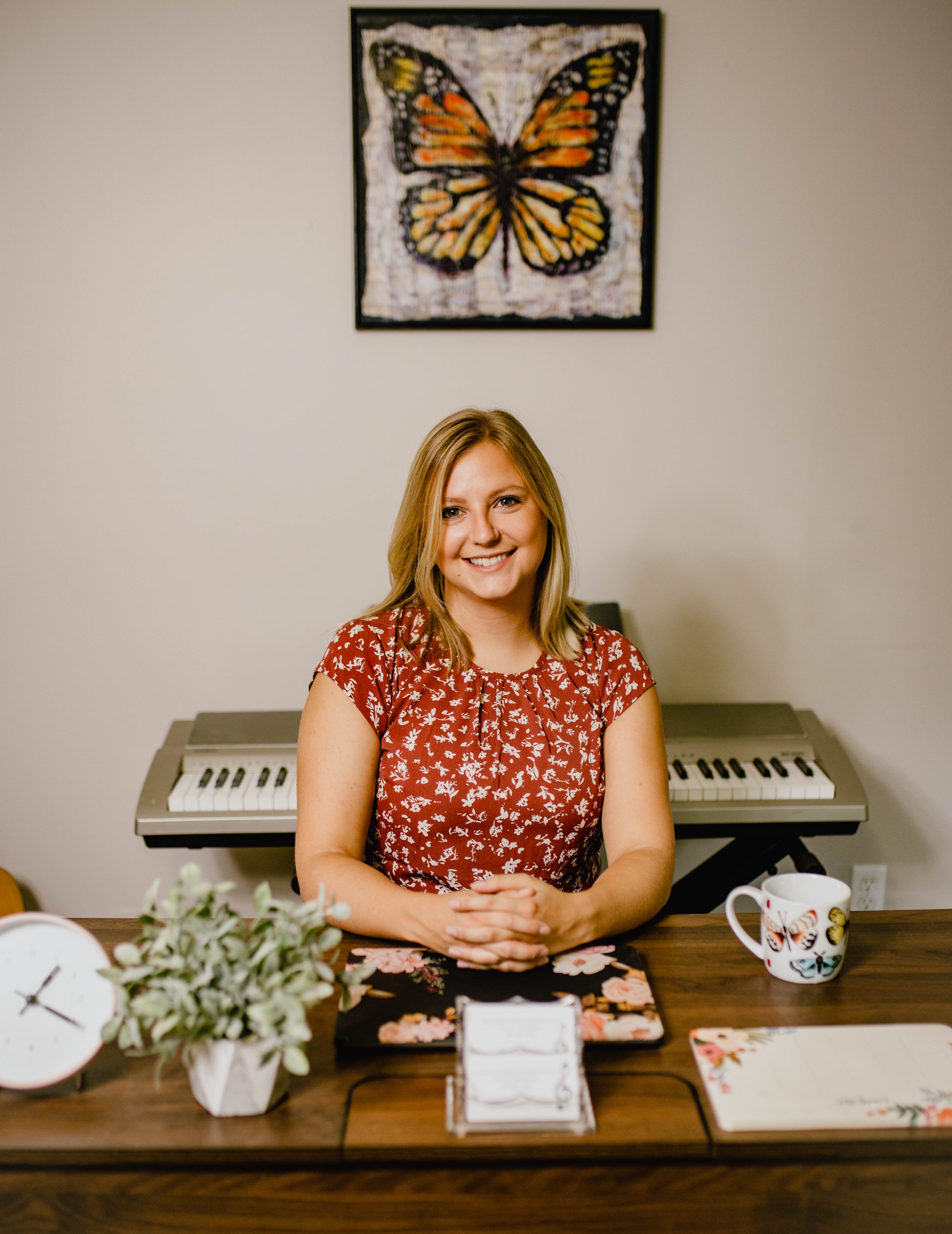A woman with blonde hair sitting at a desk with a piano in the background and a butterfly-themed artwork on the wall behind her. She is smiling, wearing a red floral dress, with a plant, a mug, and a notepad on the desk. Seattle professional head sho