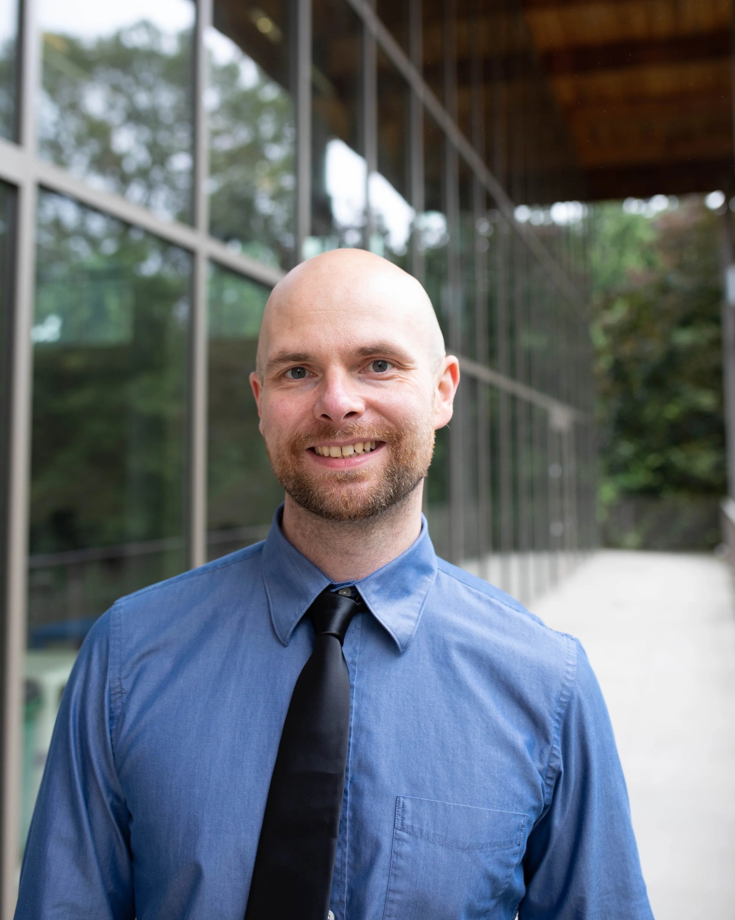 A smiling man with a beard and shaved head wearing a blue dress shirt and black tie, standing outdoors near a modern glass building with trees in the background. Seattle professional head shot photography