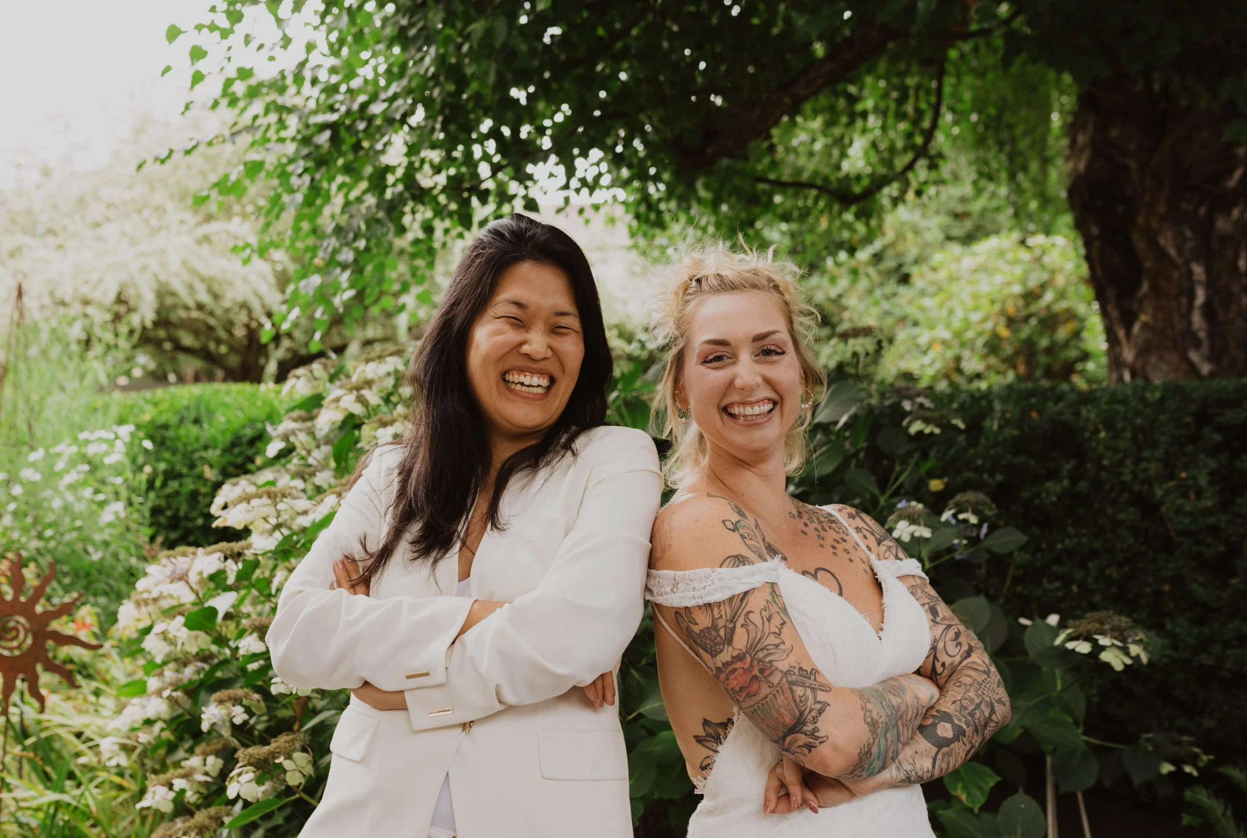 Two women smiling with arms crossed in a lush garden, one with dark hair in a white suit and the other with tattoos and a white dress. Seattle, WA wedding photography.