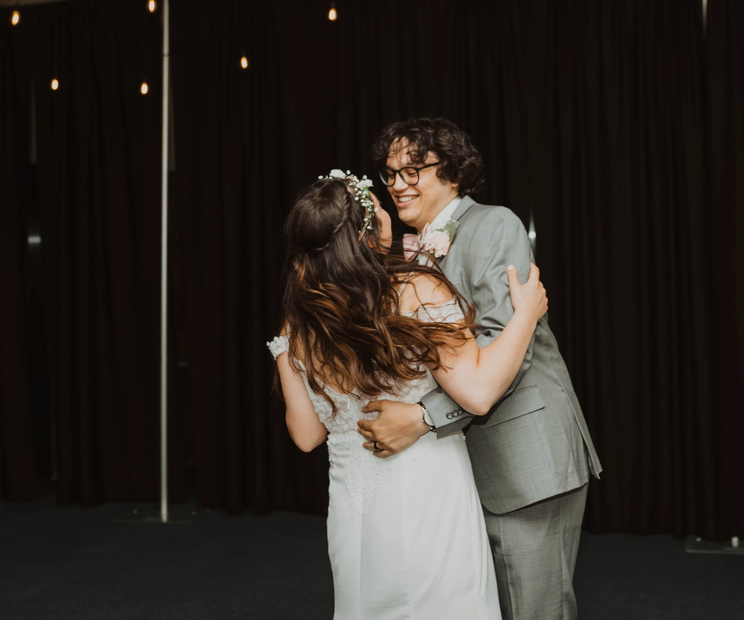 A couple dancing at their wedding, smiling and embracing each other, with dark curtains in the background. Seattle, WA wedding photography.