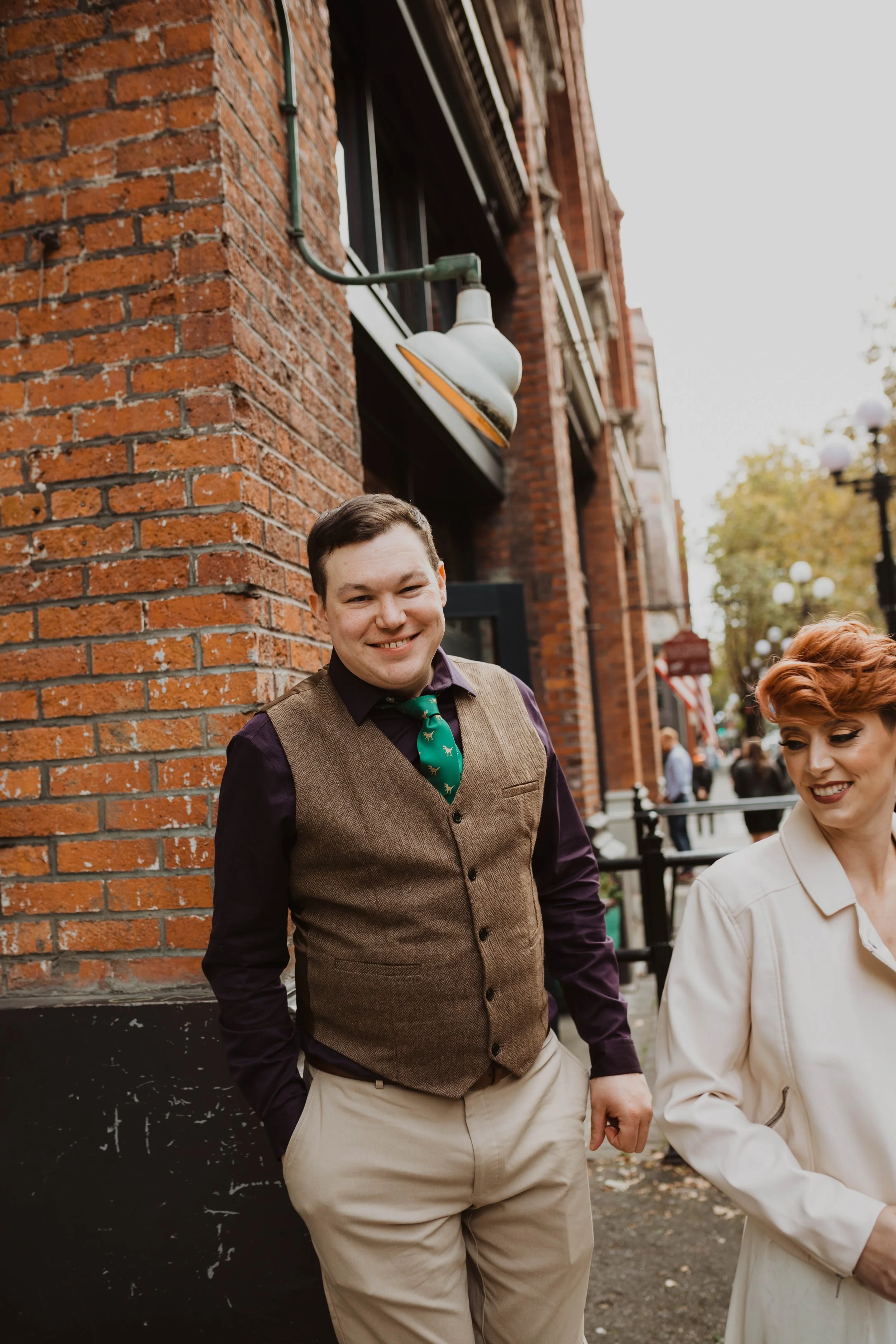 A smiling man in a brown vest, purple shirt, green tie with patterns, and beige pants standing on a city sidewalk near a brick building with a white outdoor light fixture. Next to him is a woman with short red hair in a light-colored coat, smiling an