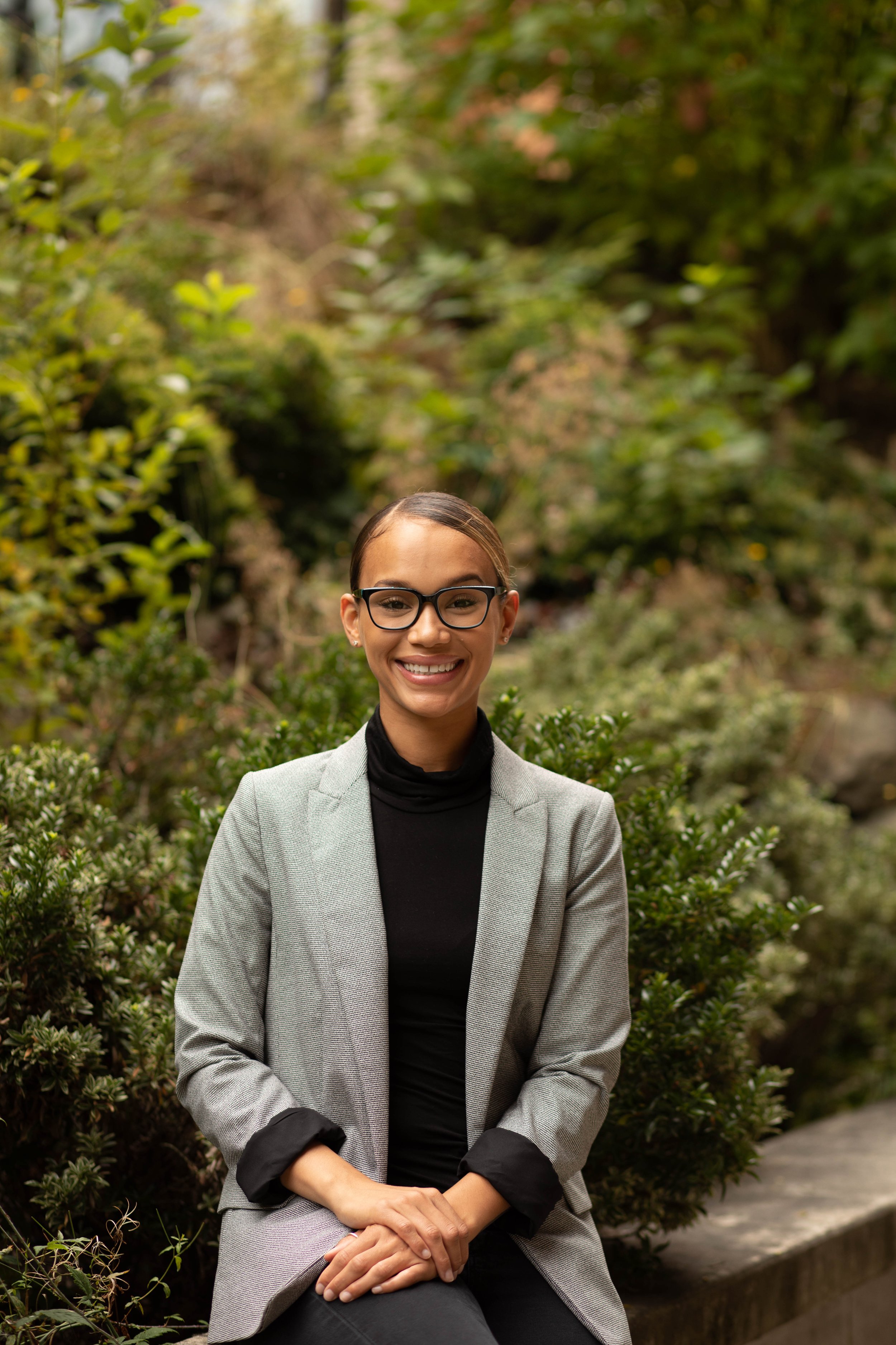 A woman in a gray blazer and black turtleneck sitting on a park bench surrounded by green bushes. Seattle professional head shot photography