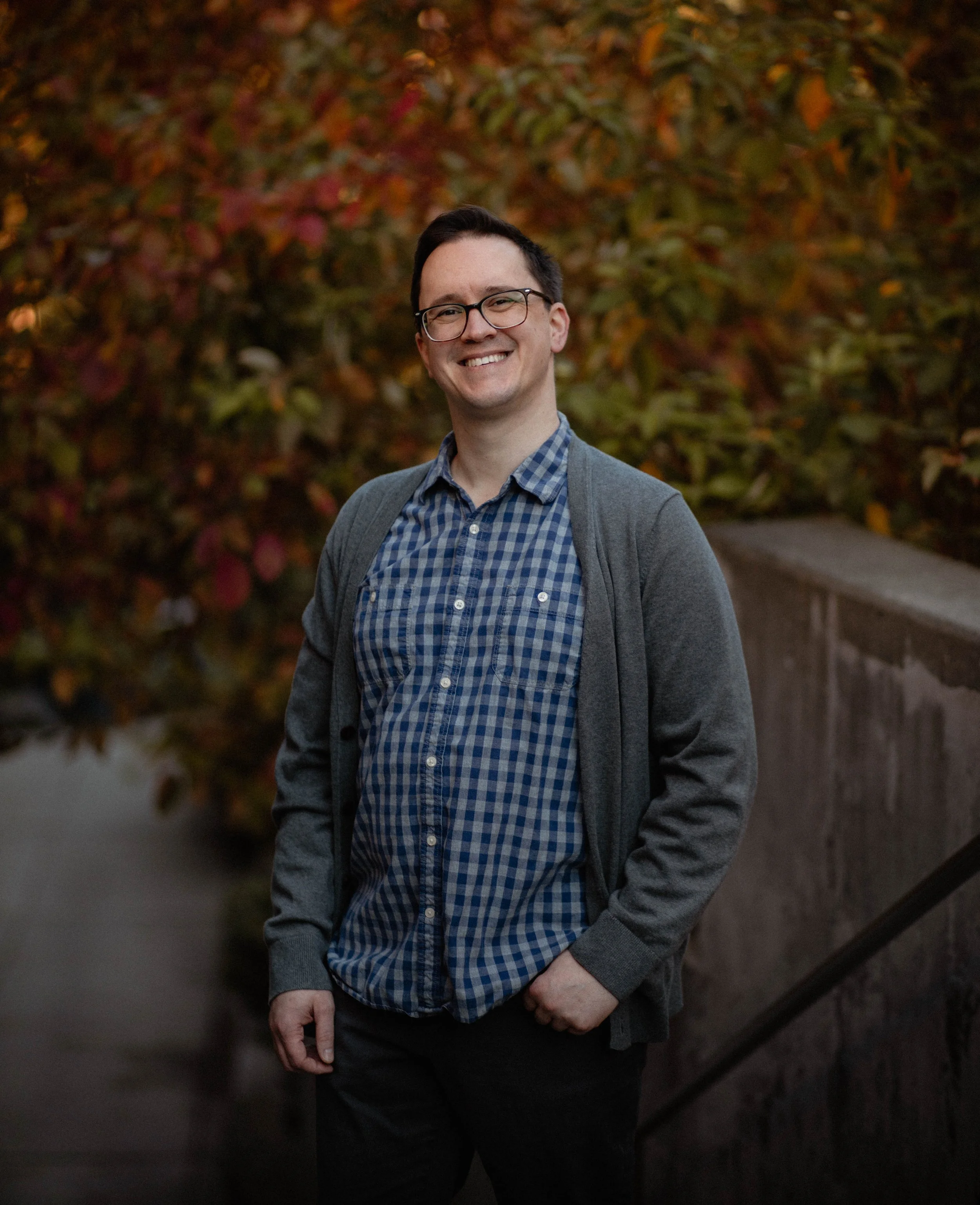 A smiling man with glasses in a checkered shirt and gray cardigan standing outdoors in front of colorful fall foliage. Seattle professional head shot photography