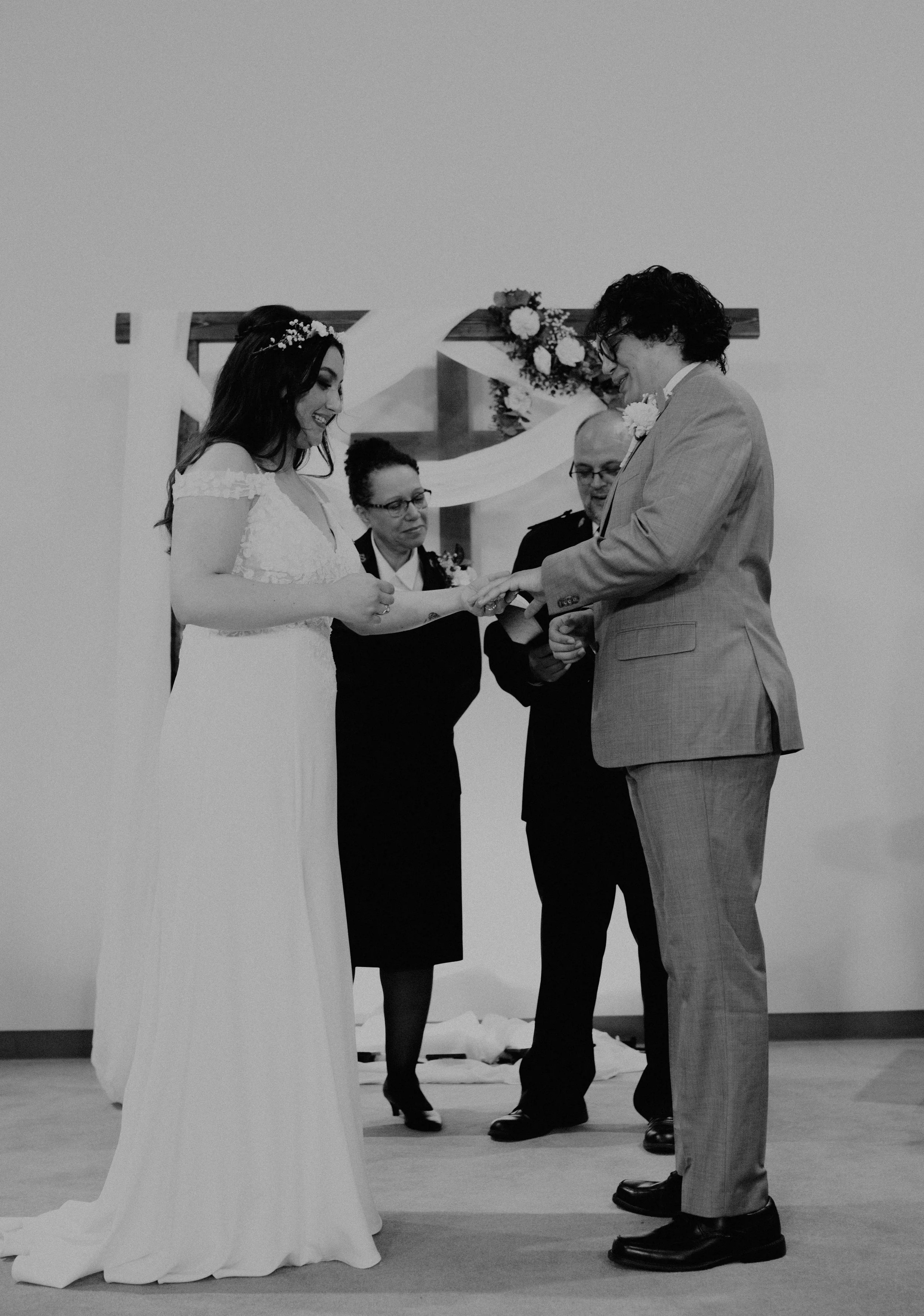 Black and white photo of a wedding ceremony, showing a bride and groom exchanging rings with officiant and a witness present. Seattle, WA wedding photography.