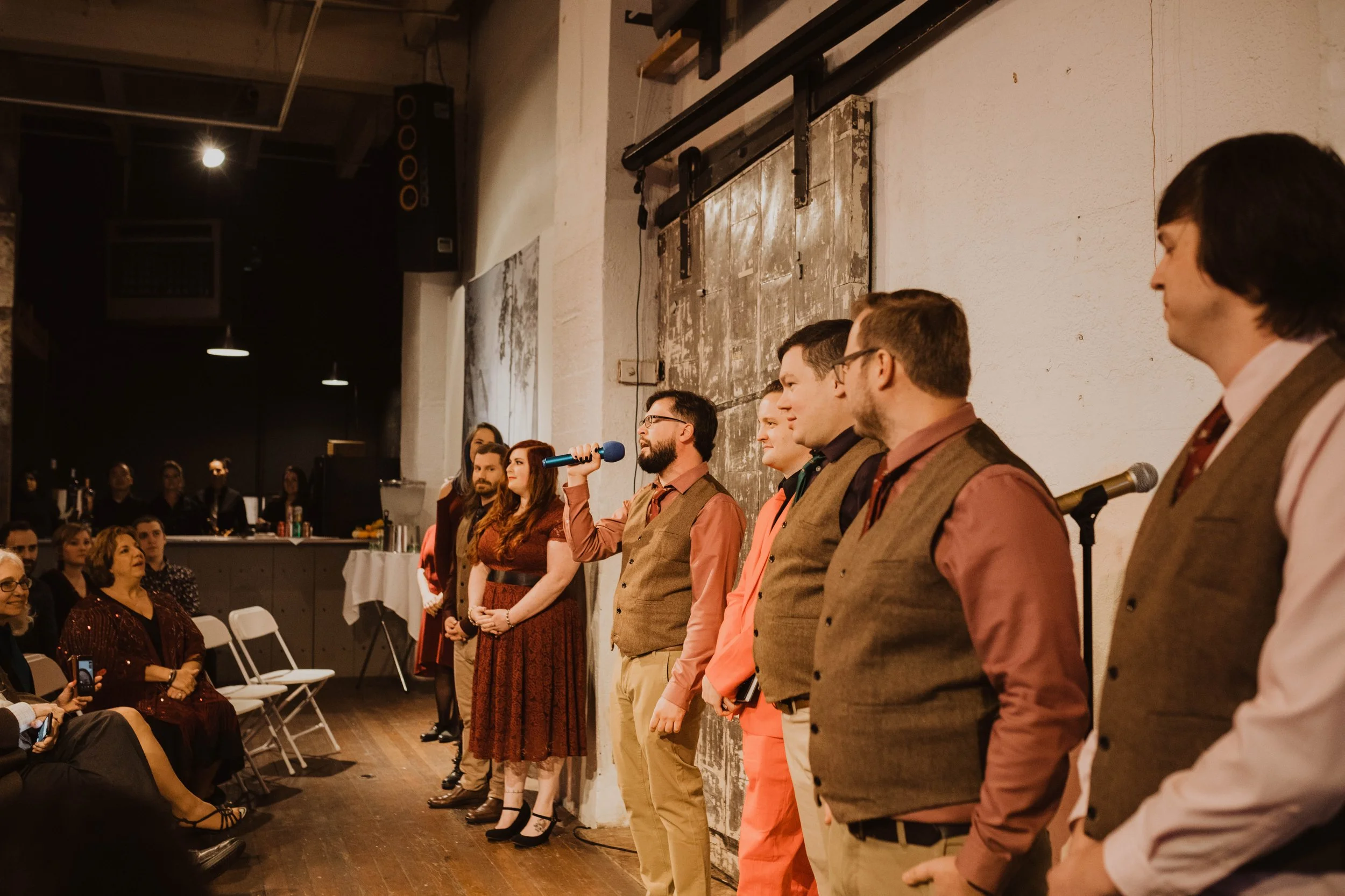 A group of people standing on a stage in front of an audience, with one person holding a microphone. The setting appears to be an indoor event or performance, with a casual and artistic atmosphere. Pioneer Square, Seattle, WA wedding photography.