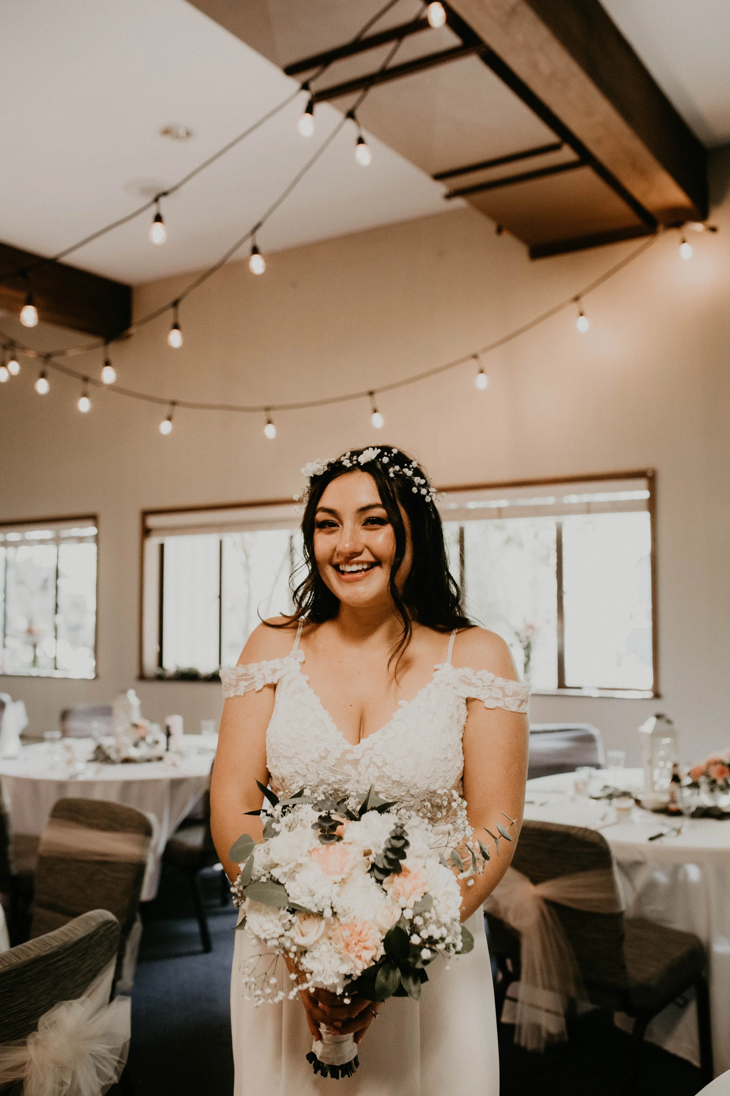 A bride holding a bouquet in a decorated reception hall with string lights, smiling. Seattle, WA wedding photography.