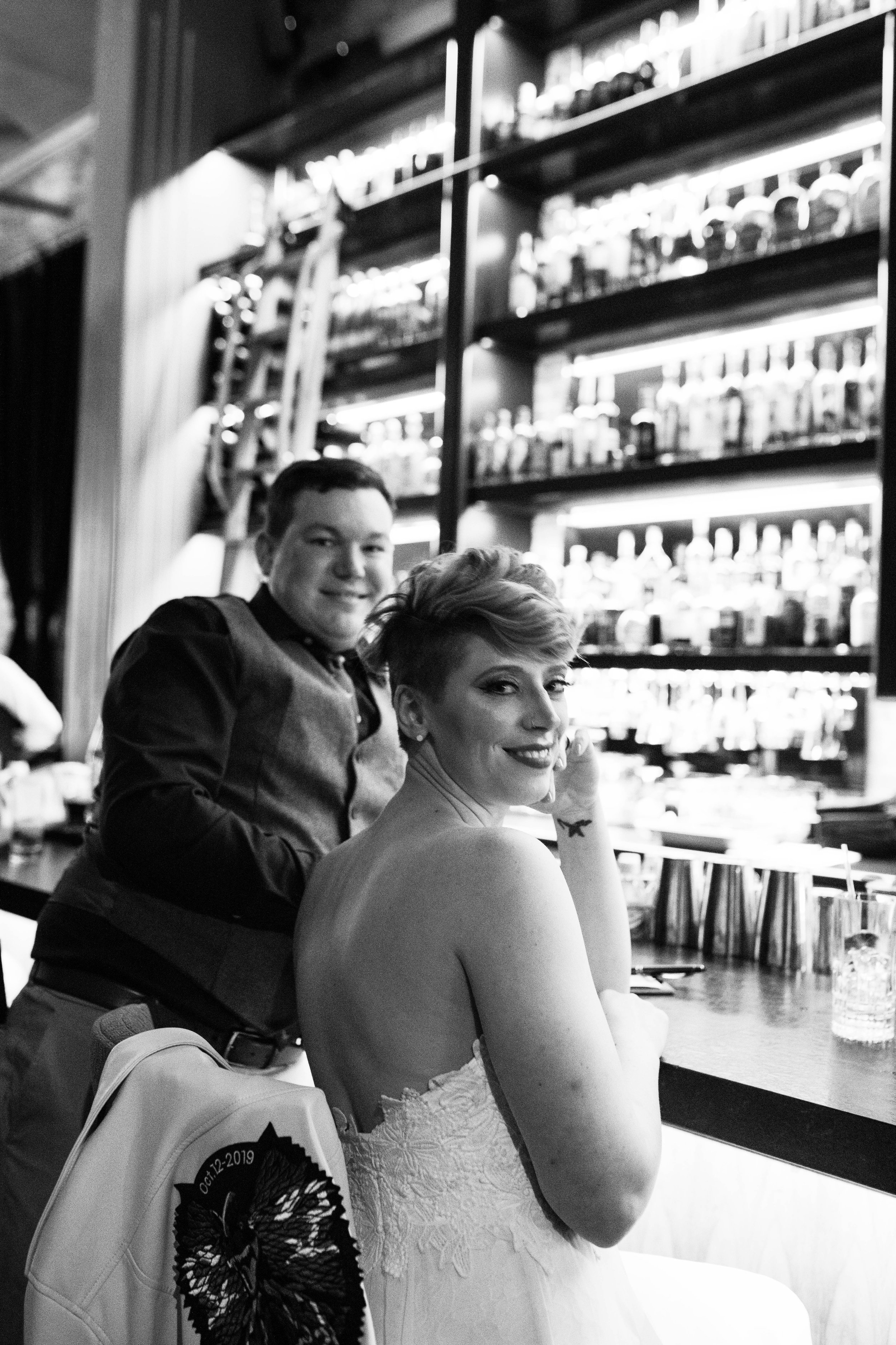 Two people at a bar, one woman in a strapless dress and one man in a shirt and vest, with a well-stocked bar in the background. Pioneer Square, Seattle, WA wedding photography.