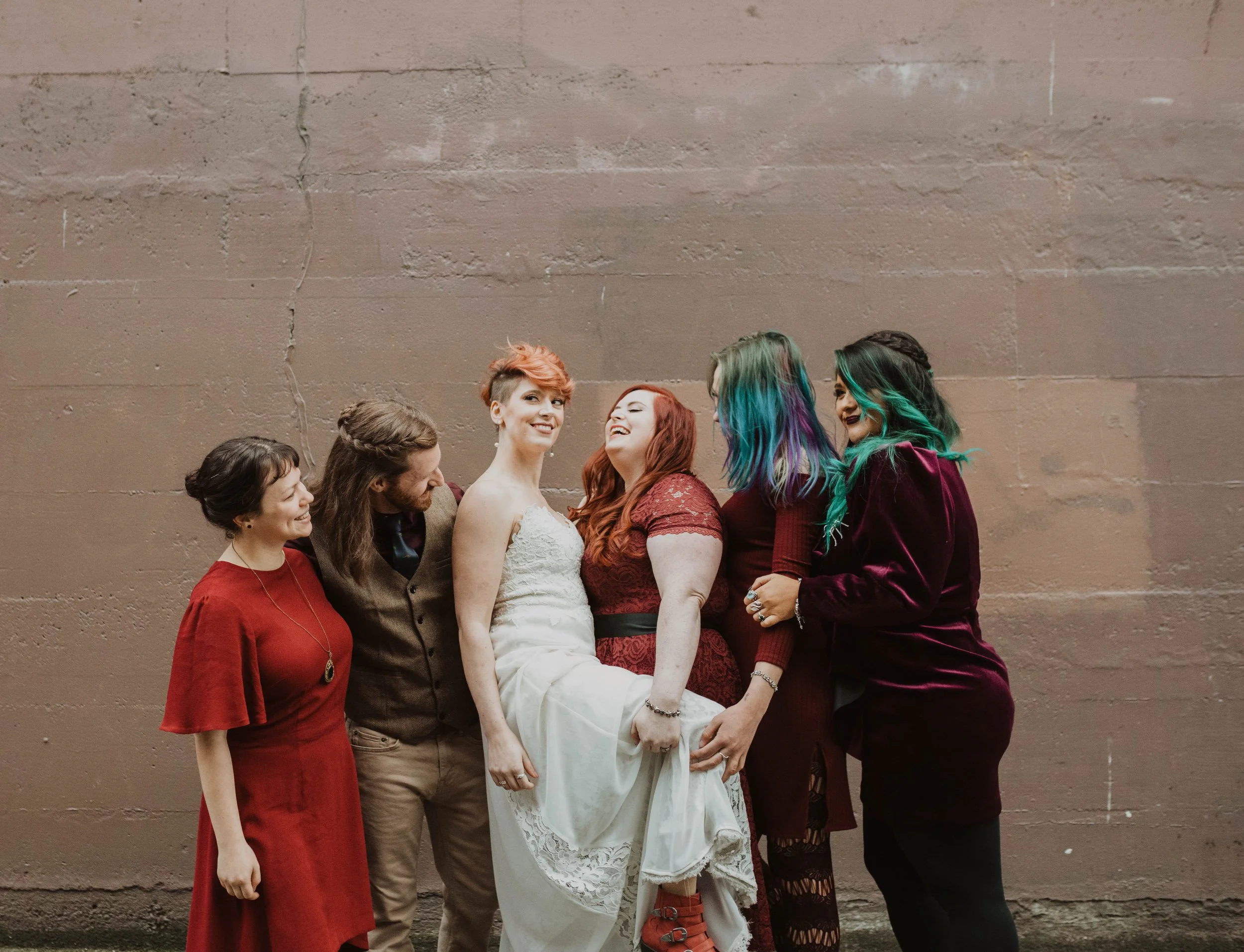A group of six friends standing against a plain wall, laughing and smiling, with one woman in a wedding dress at the center. The women have colorful hairstyles, and the group is dressed in a mix of casual and formal attire.
