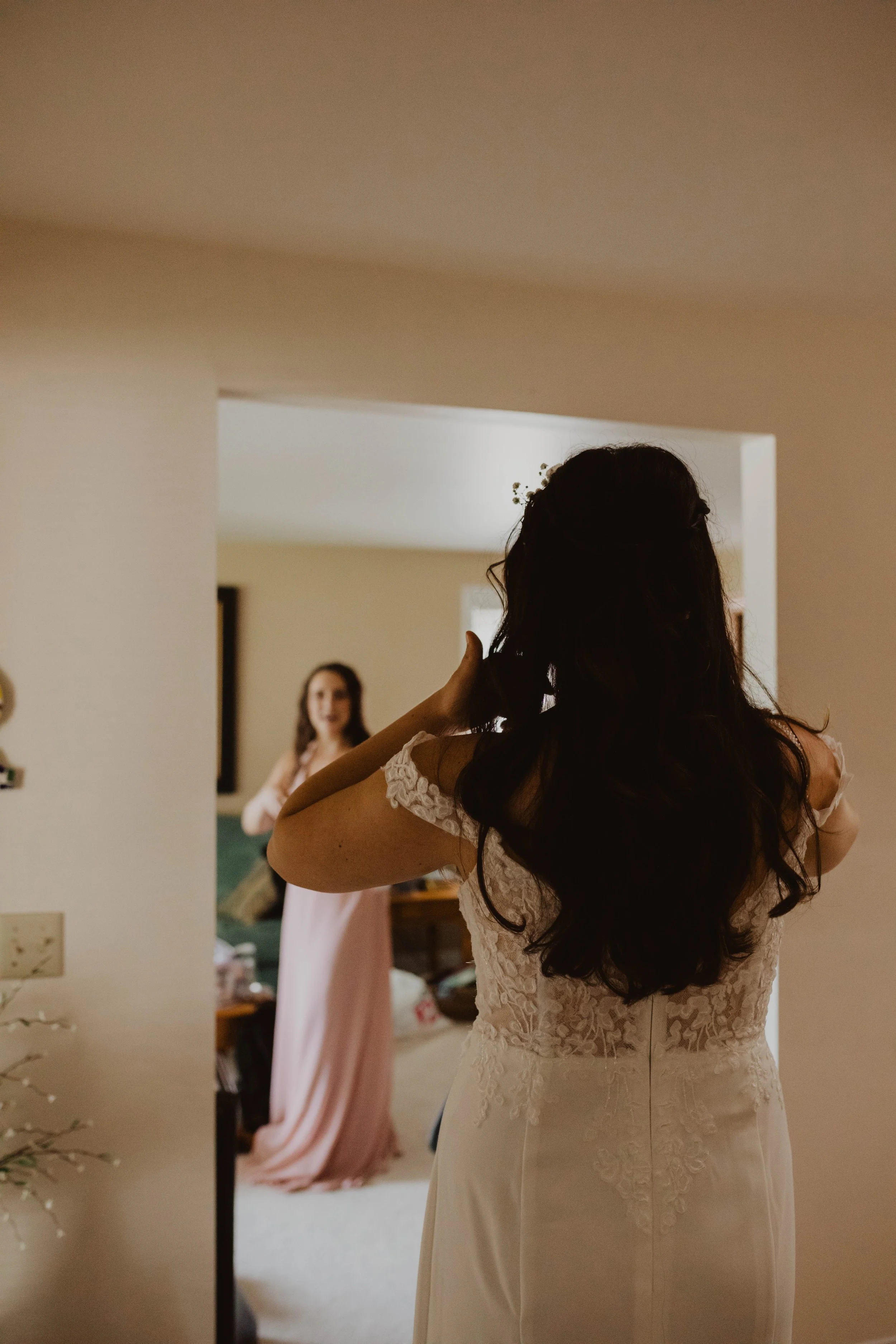 A bride with long dark hair wearing a white lace dress, adjusting her hair while looking at another woman in a pink dress in a living room. Seattle, WA wedding photography.