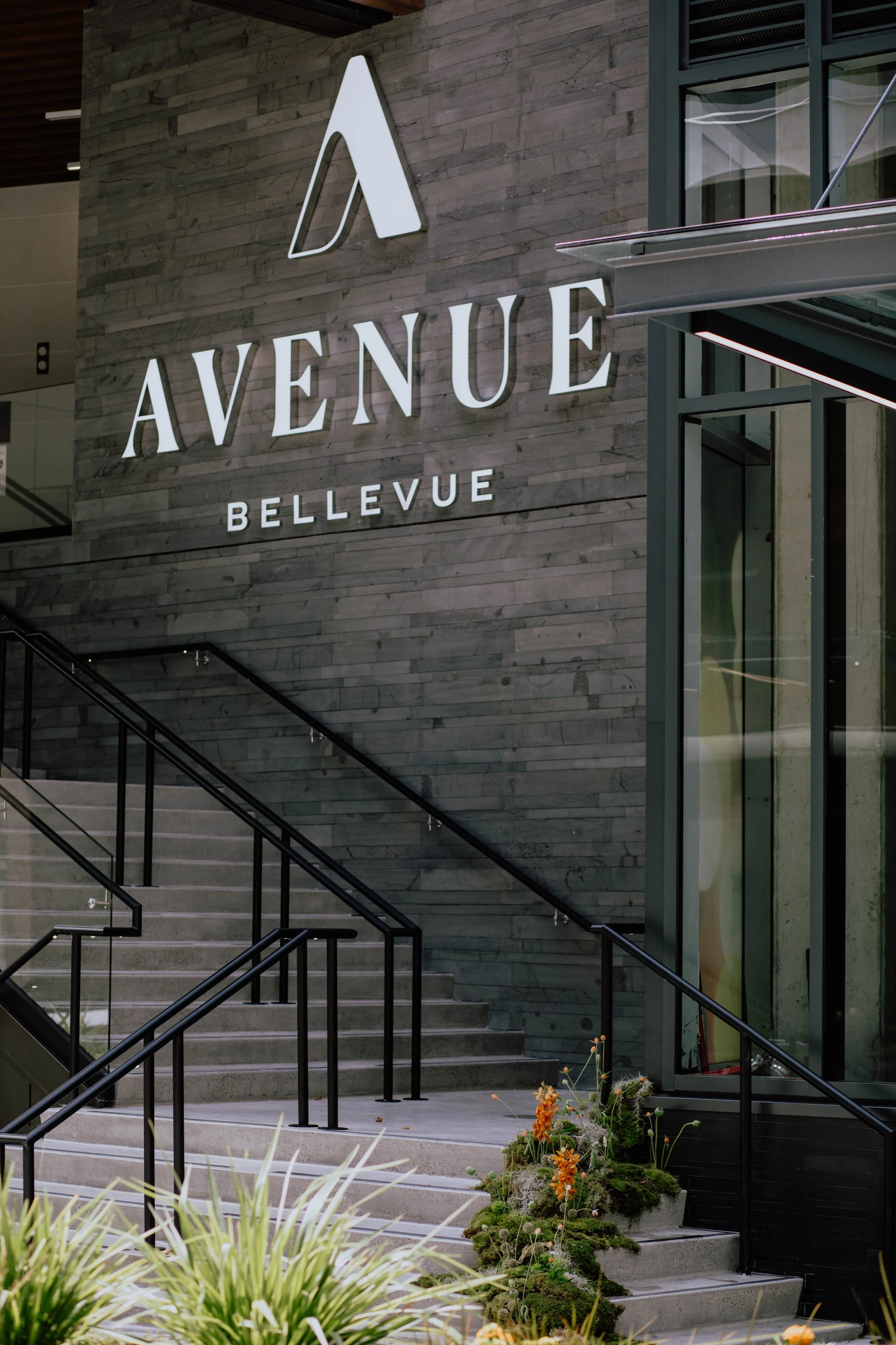 Exterior of a modern building with stairs, black railings, and a large sign reading 'Avenue Bellevue' on a gray stone wall. Glass windows reflect the surroundings, and there are plants and flowers at the base of the stairs.