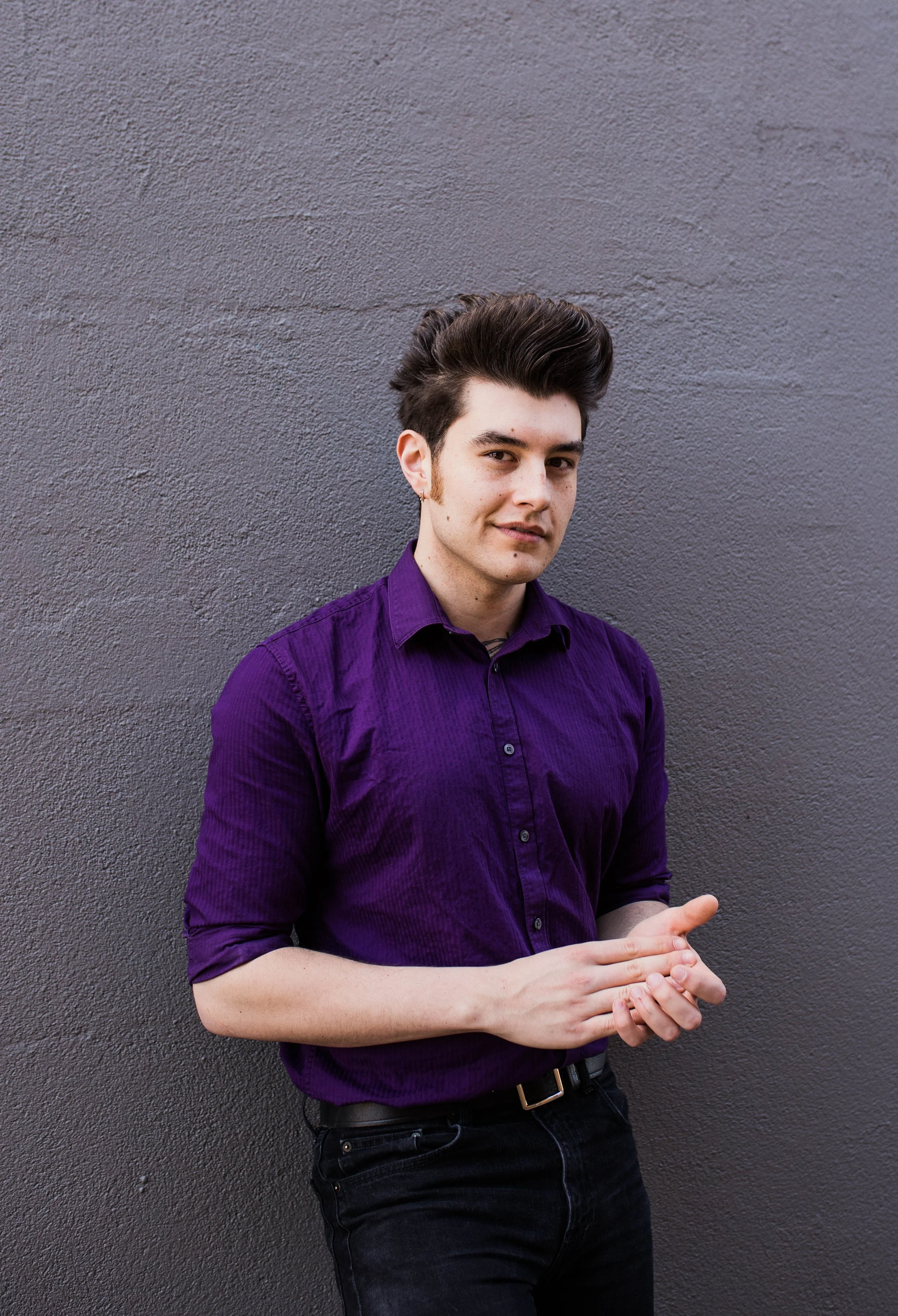 A young man with dark, styled hair, wearing a purple button-up shirt, stands against a plain gray wall, looking slightly to the camera with a subtle smile. Seattle professional head shot photography