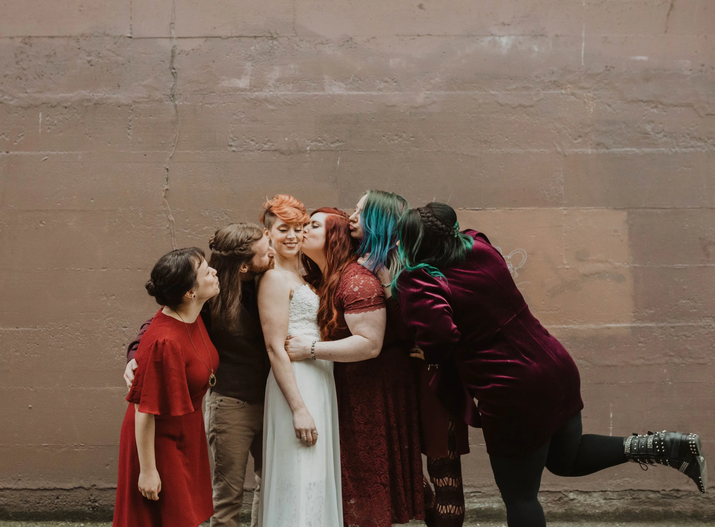 Group of friends and family gather closely together, with one woman receiving kisses on the cheek, against a plain brick wall, celebrating a wedding or special occasion. Pioneer Square, Seattle, WA wedding photography.