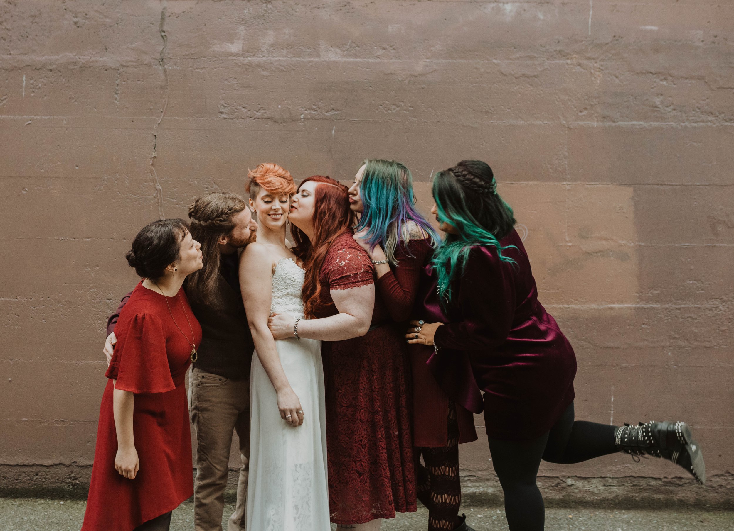 Group of six women and one man standing against a plain brown wall, dressed casually and affectionately hugging and kissing each other, with one woman lifting one leg slightly off the ground. Pioneer Square, Seattle, WA wedding photography.
